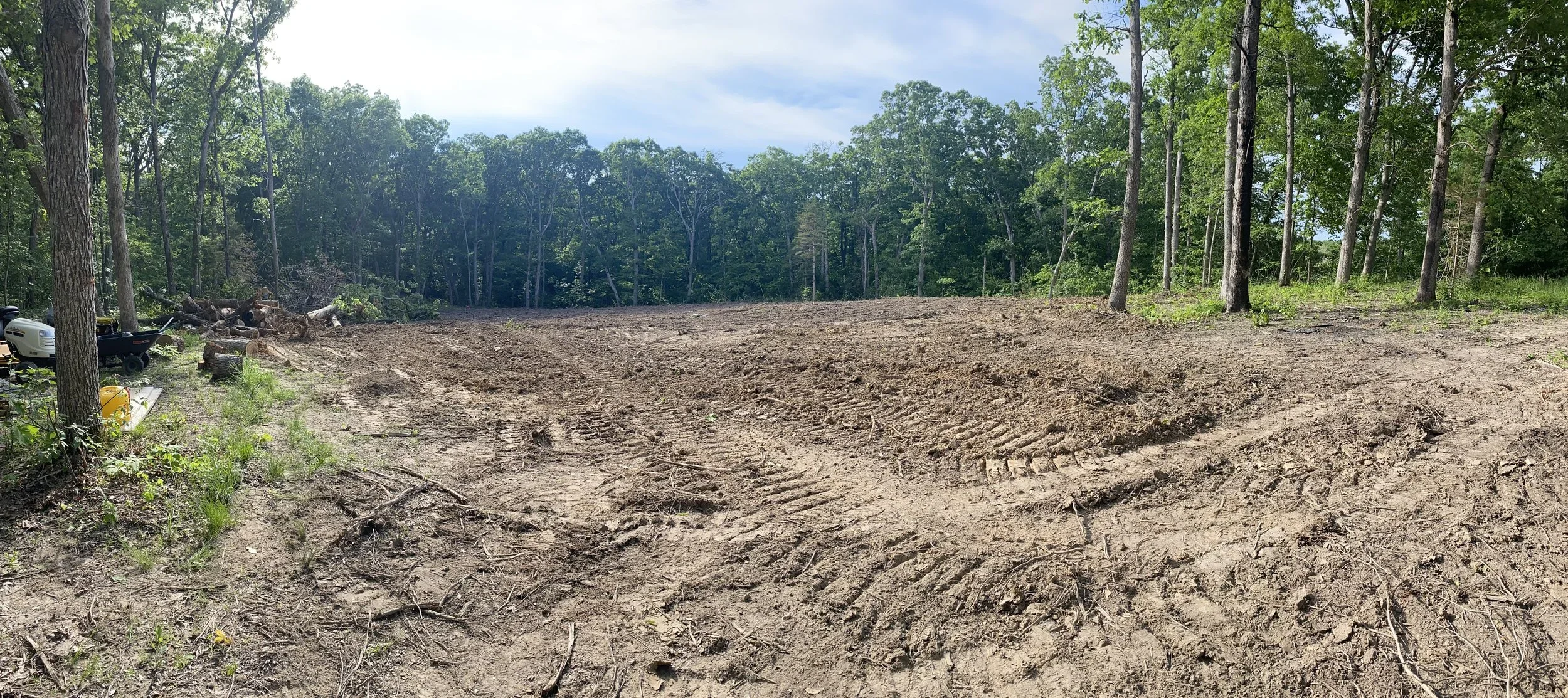 Cleared land in a wooded area with trees in the background and construction equipment on the left side.