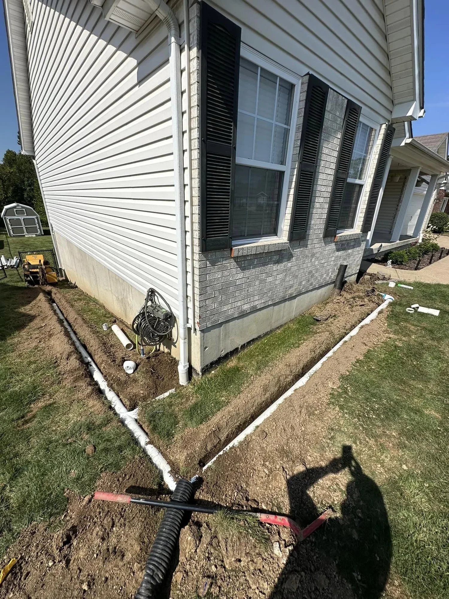 Installation of underground water drainage pipes along the side of a house with vinyl siding and brick exterior.