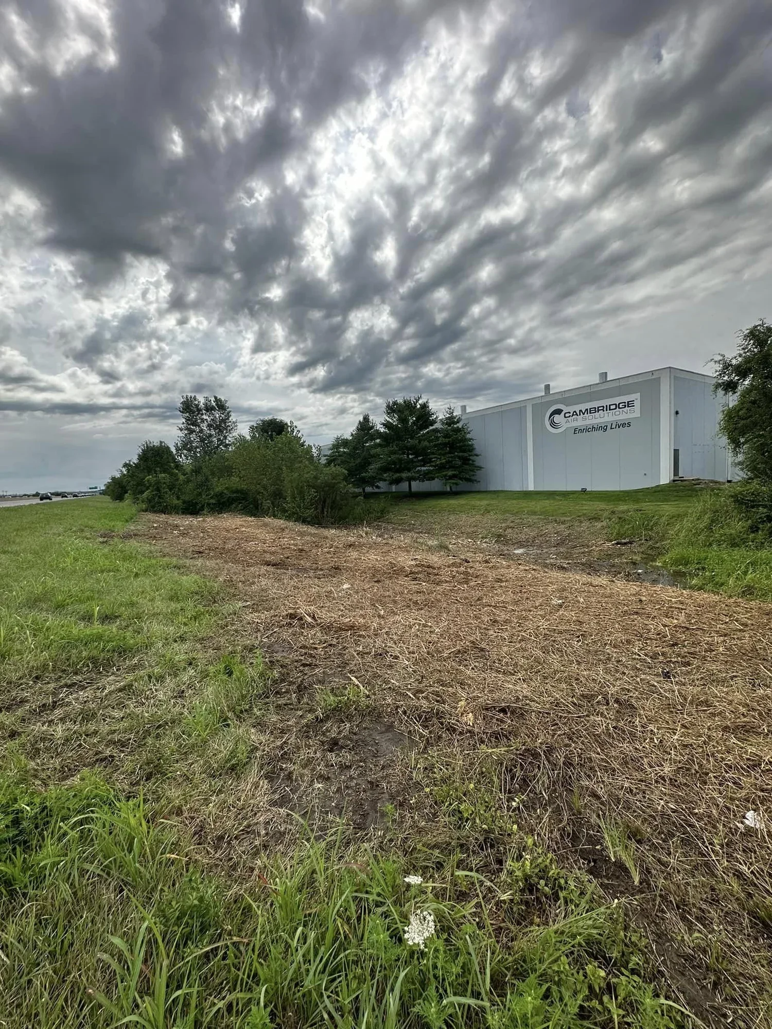 Industrial building with a sign that reads 'Cambridge Air Solutions' and 'Enriching Lives' on a cloudy day, surrounded by green grass and trees.