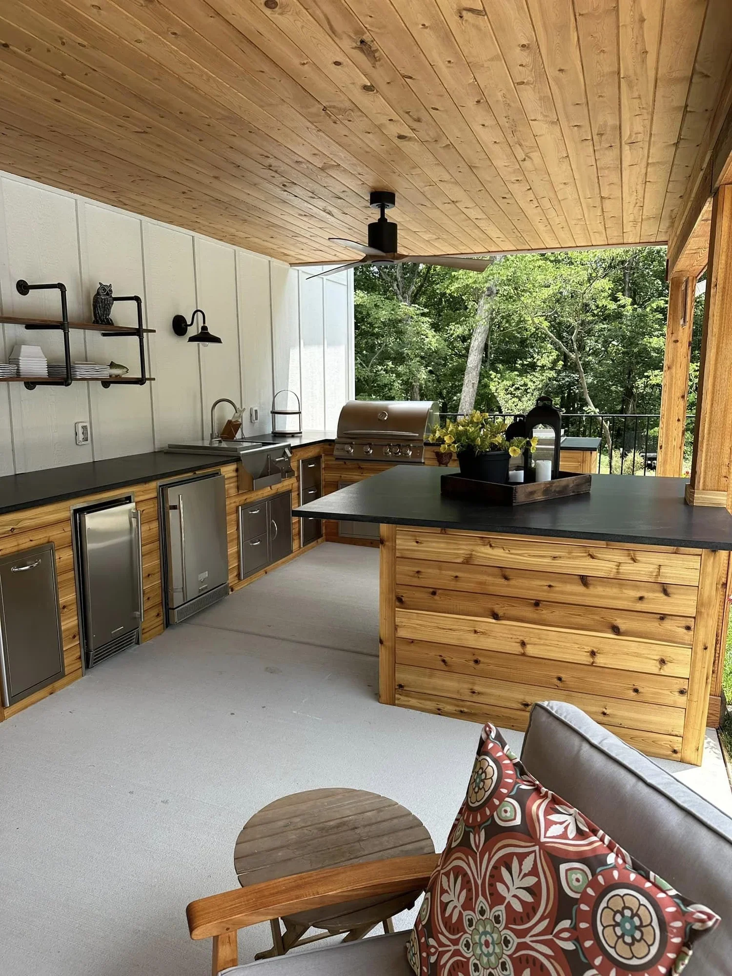 A backyard outdoor kitchen area with a wooden ceiling, a ceiling fan, a black countertop with a built-in grill, metal cabinets, and a view of green trees outside. There is a small round wooden table and a chair with a floral-patterned pillow.