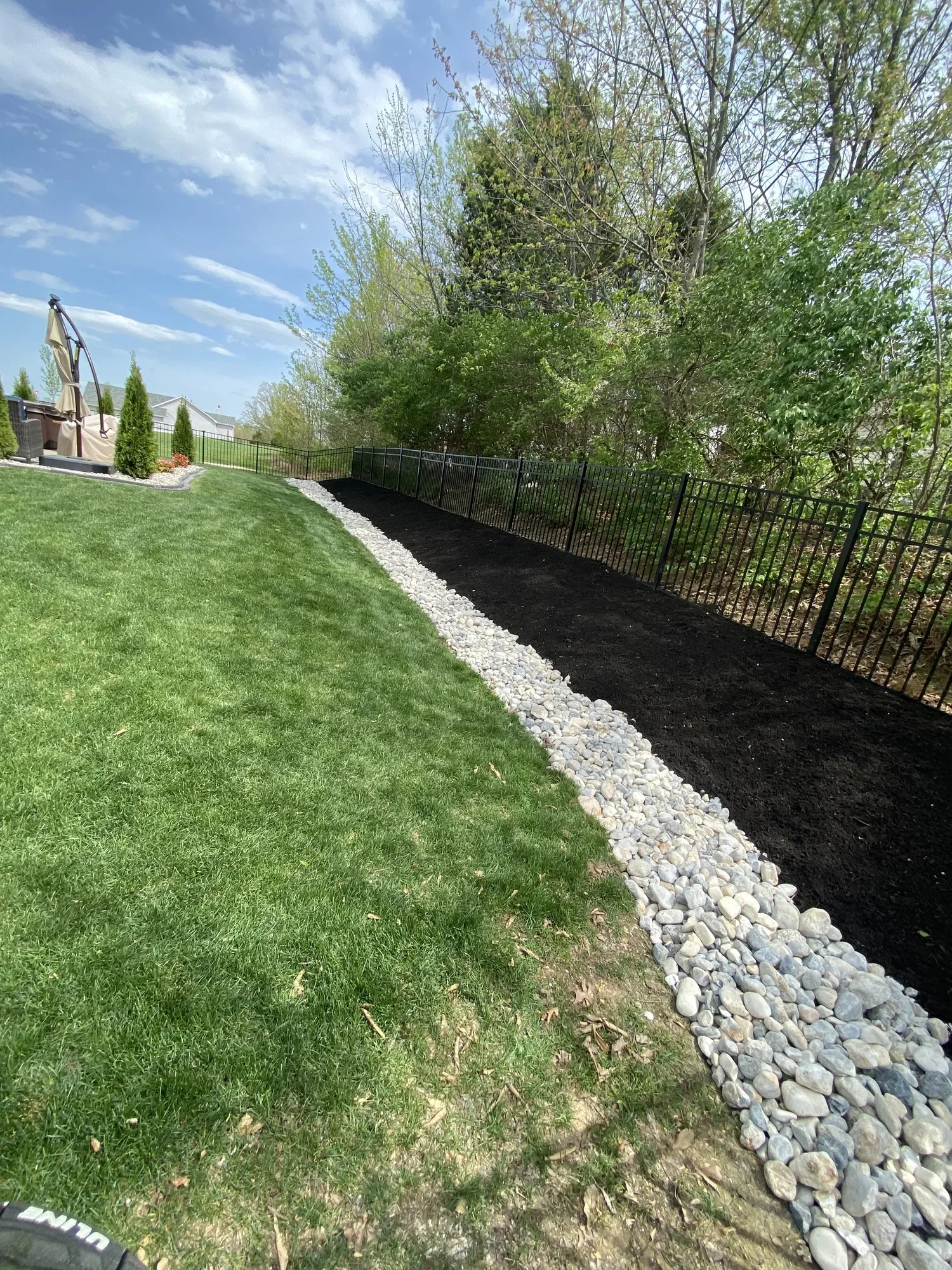 A backyard with a well-maintained lawn, a black metal fence, a freshly laid black mulch garden bed with white river rocks along the edge, and trees with fresh green leaves under a partly cloudy sky.