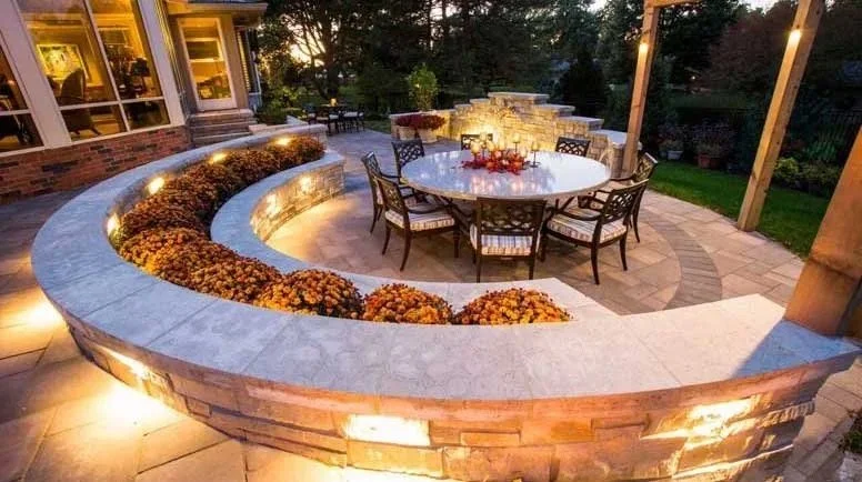 Outdoor patio area with a circular dining table surrounded by six chairs, a curved stone wall with built-in lighting, flower beds with orange and yellow flowers, and string lights overhead, in a backyard at dusk.