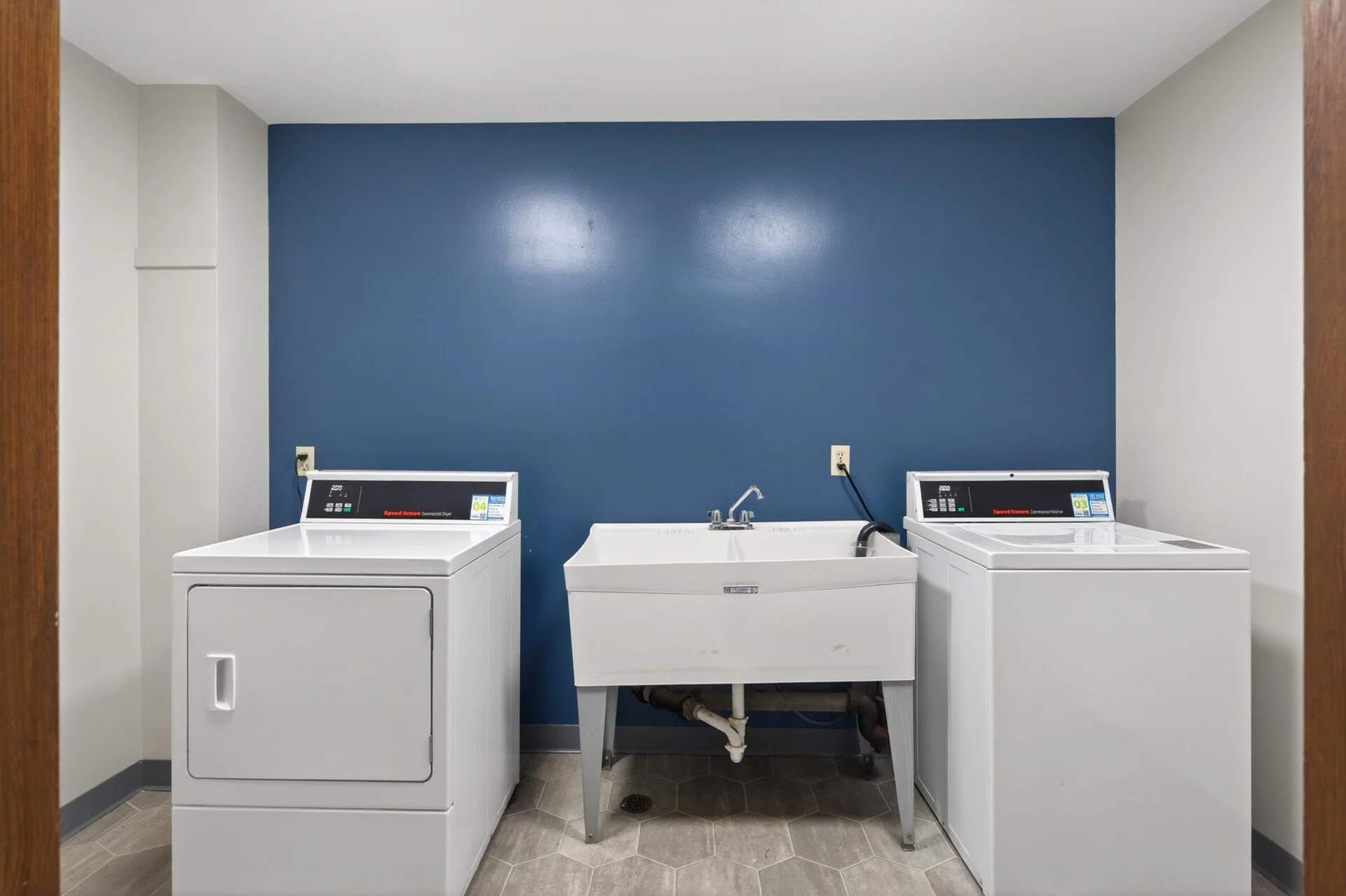 A laundry room with a blue accent wall, two white washing machines, a white utility sink, and a dark pipe under the sink at Imperial Heights in West St. Paul, MN