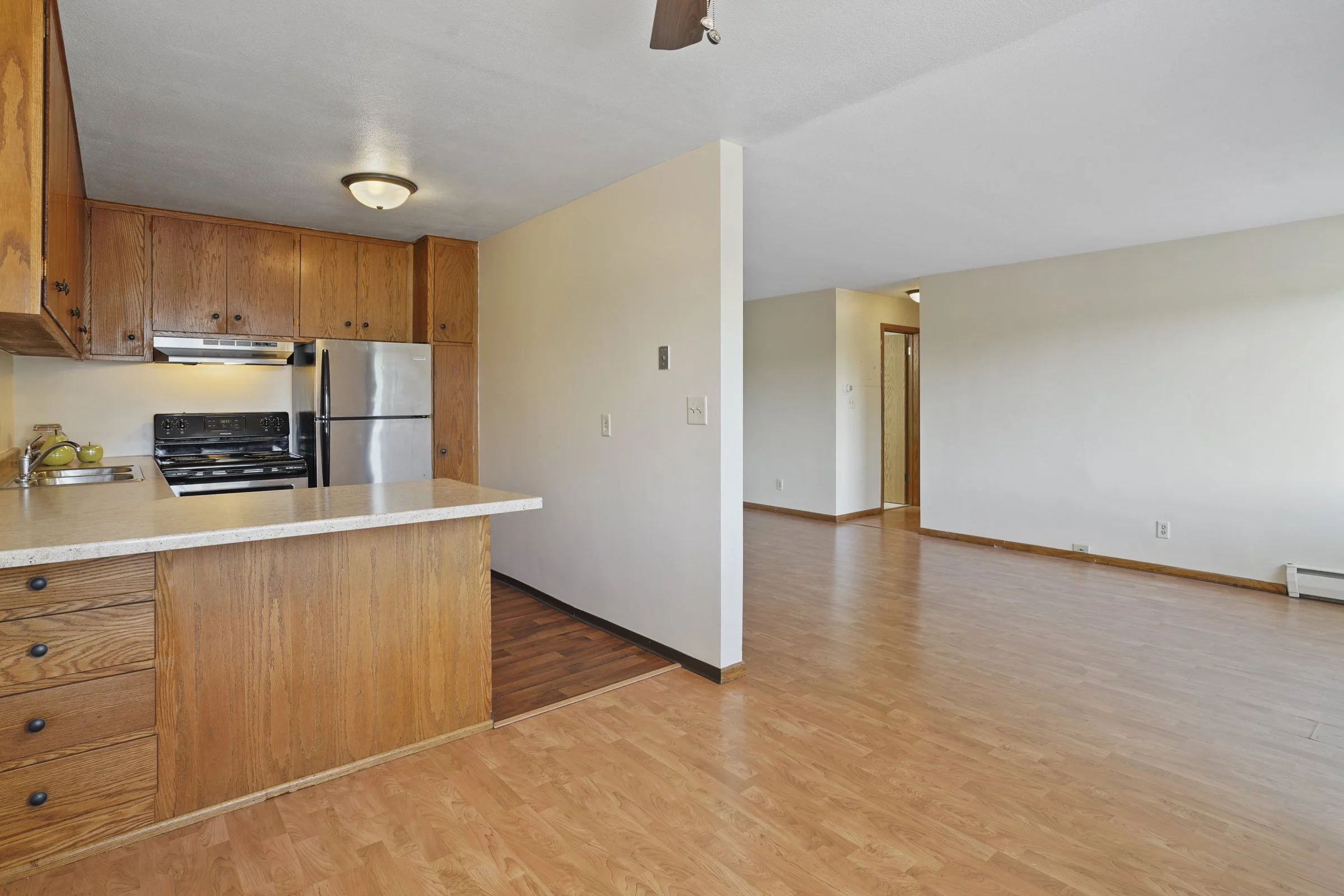 Inside view of an empty living room with wooden floors, white walls, a kitchen with wooden cabinets, a yellowish countertop, and a stainless steel refrigerator. The ceiling has a flush mount light fixture and a ceiling fan is partially visible. There