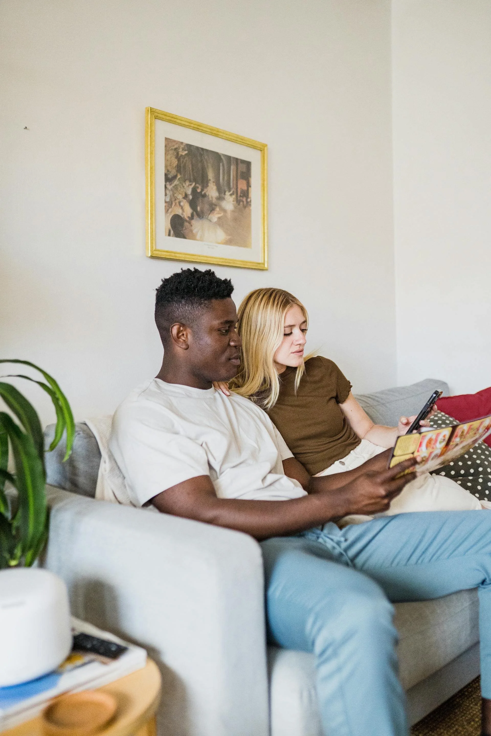 A young man with dark skin and short curly hair, and a young woman with light skin and long blonde hair, sitting on a beige couch in a living room, looking at a smartphone. There is a framed painting on the wall behind them.