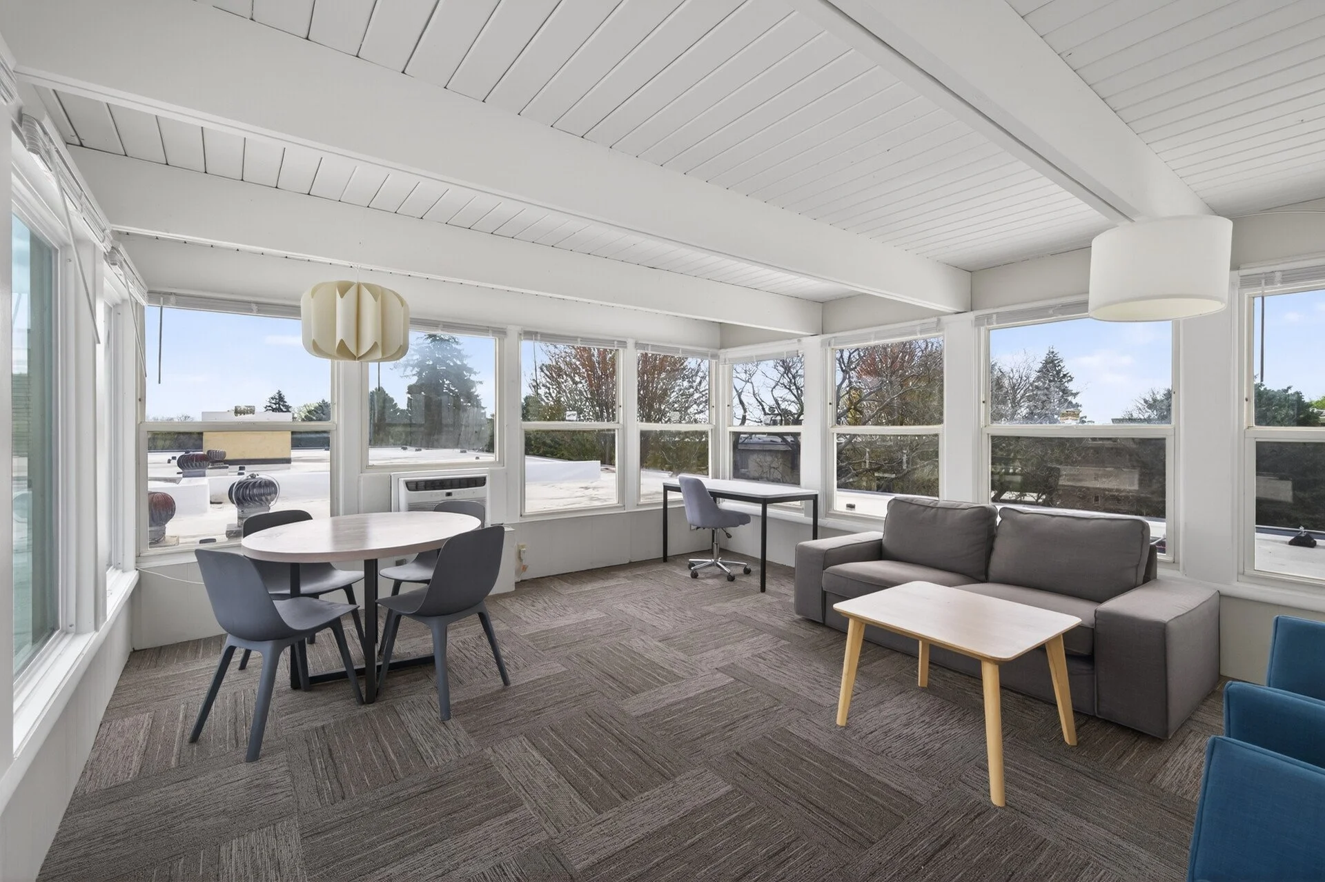 Sunroom with large windows, gray sofa, round dining table with four chairs, small desk with office chair, ceiling lights, and view of trees outside at Imperial Heights in West St. Paul, MN