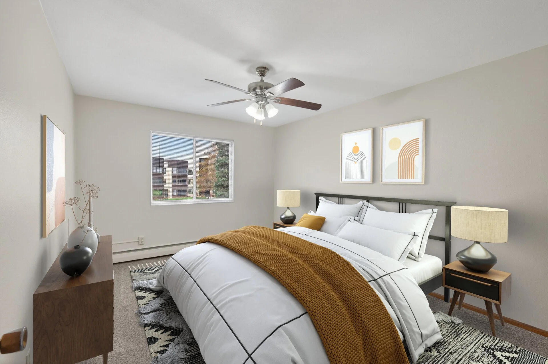 A bedroom with a bed, mustard-colored blanket, and pillows, two nightstands with lamps, a window with blinds, and two framed geometric art pieces on the wall at Imperial Heights in West St. Paul, MN