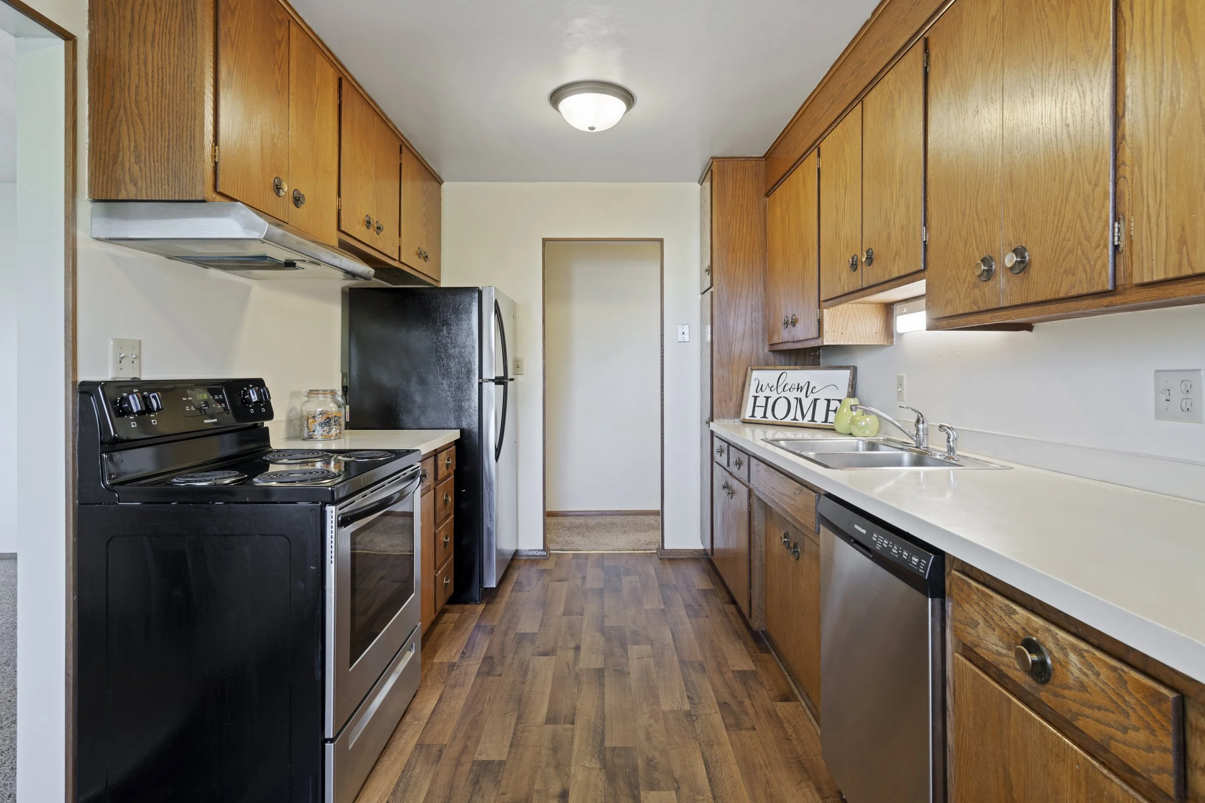 A kitchen with wooden cabinets, black appliances, and a white countertop, featuring a welcome home sign and two green decorative vases near the sink.