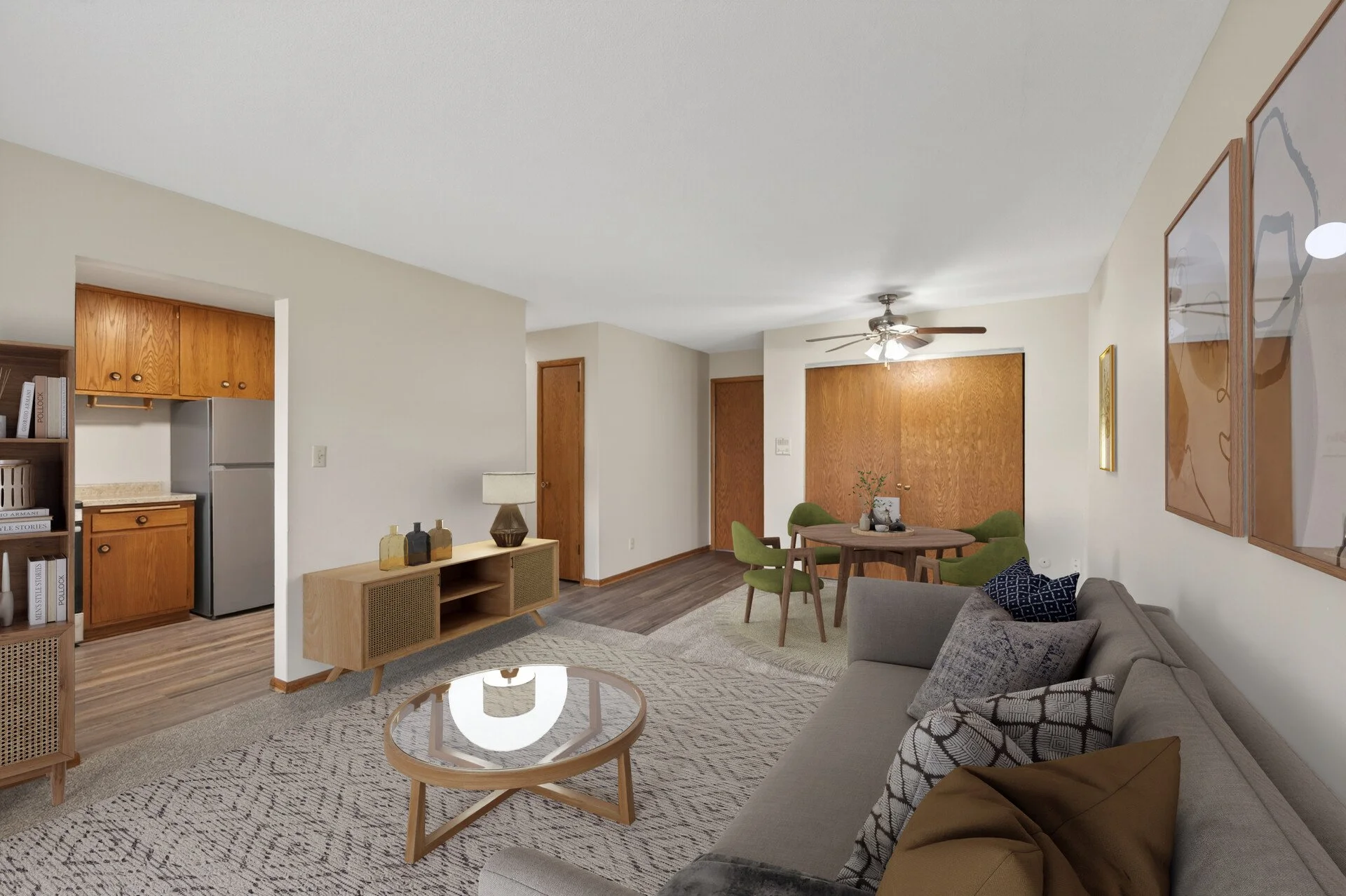 Living room with beige walls, hardwood floors, a gray sofa with pillows, a wooden coffee table, a small dining table with green chairs, a ceiling fan, wall art, a wooden media console, and a partial view of the kitchen with wooden cabinets and a refr