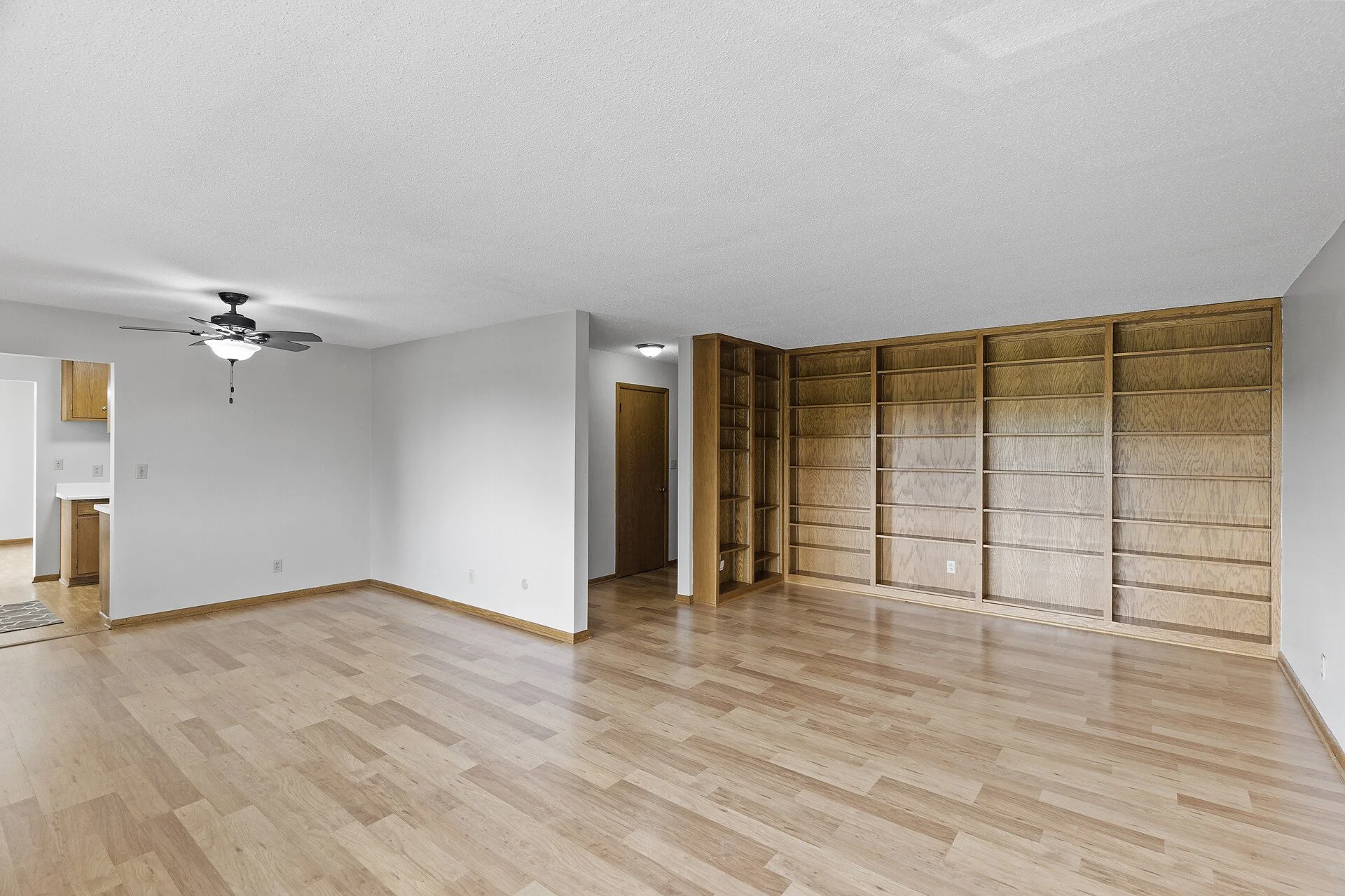 Empty living room with light wood flooring, white walls, a ceiling fan with light, and built-in wooden shelves against one wall.