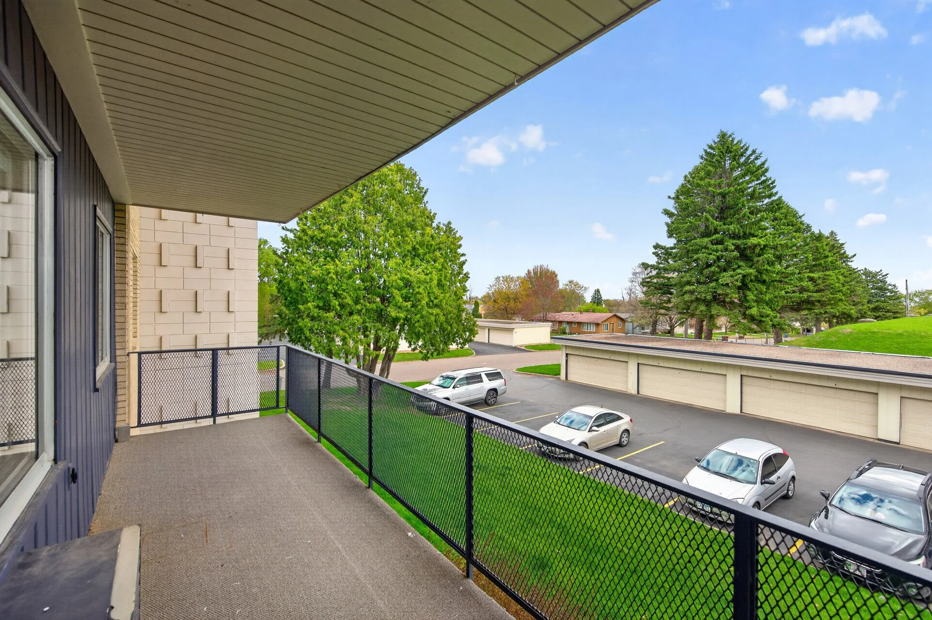 View from a balcony at Imperial Heights in West St. Paul, MN overlooking a parking lot, green trees, and residential houses under a blue sky with some clouds.