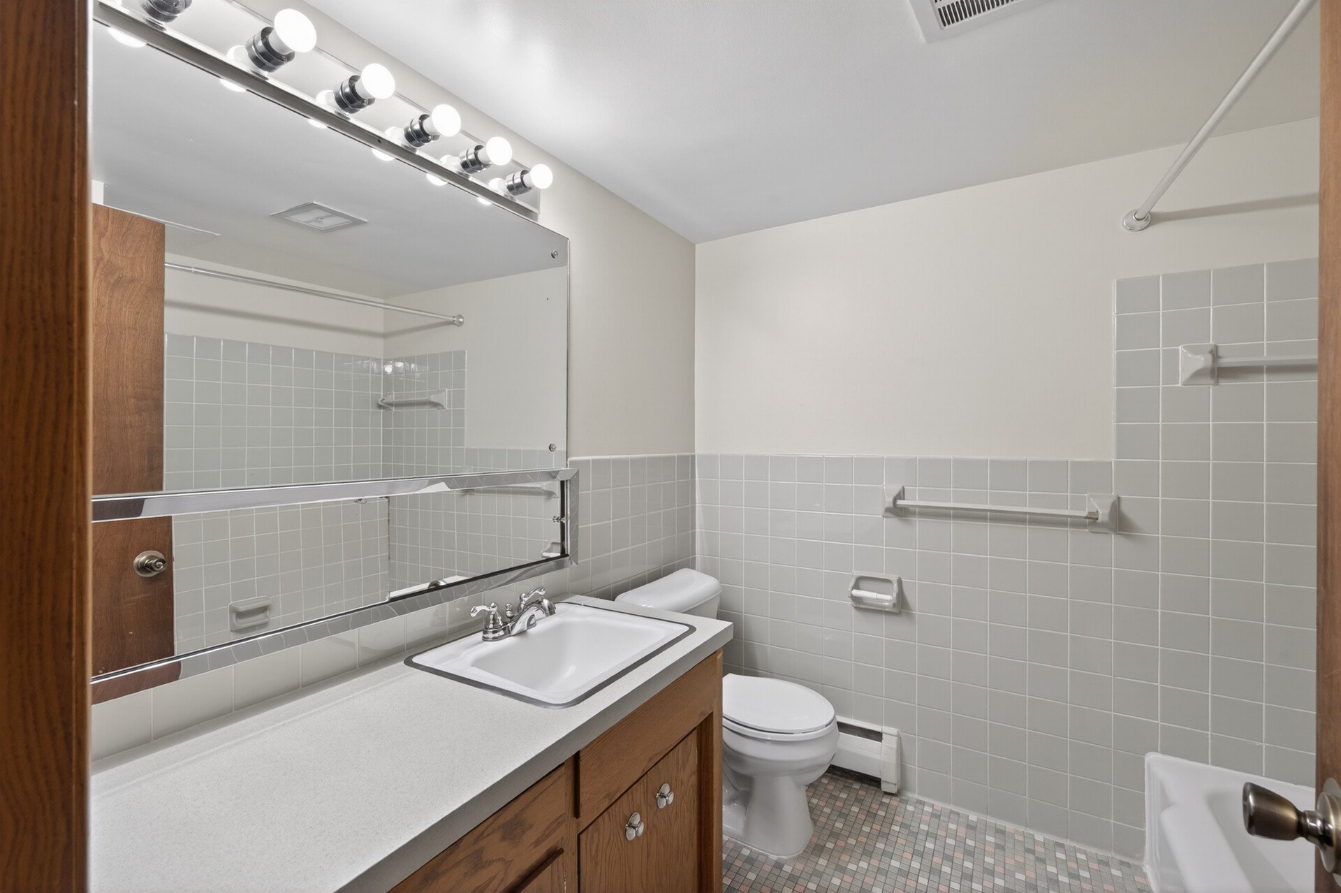 A bathroom with a white sink and countertop, wooden cabinetry, a large mirror, and white tiled walls. There is a toilet, a towel rack, and a tiled floor at Imperial Heights in West St. Paul, MN