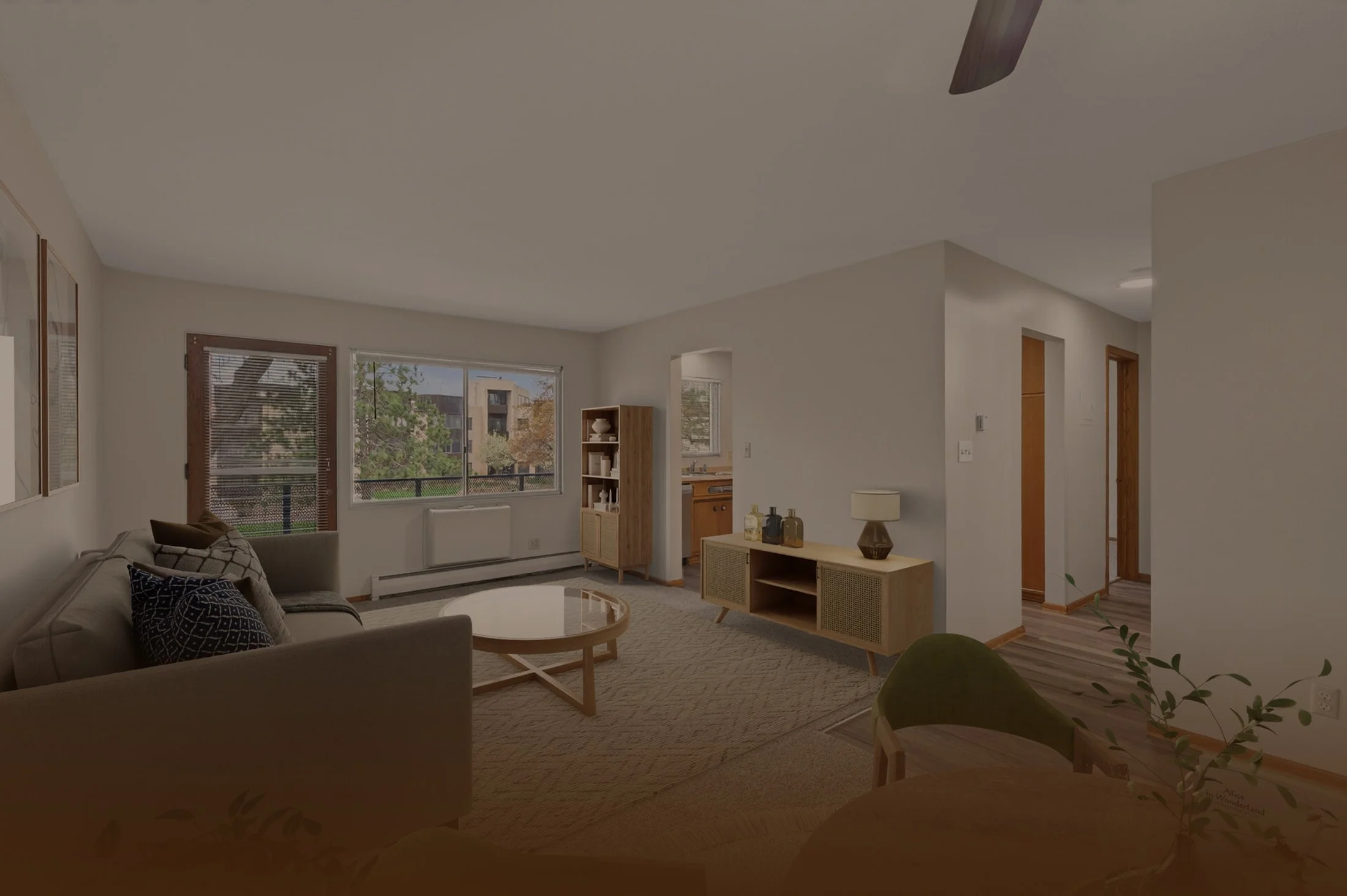 A living room with beige walls and ceiling, large windows with blinds showing trees outside, a grey couch with patterned pillows, a round coffee table, a wooden shelving unit, and decorative items like lamps and vases.
