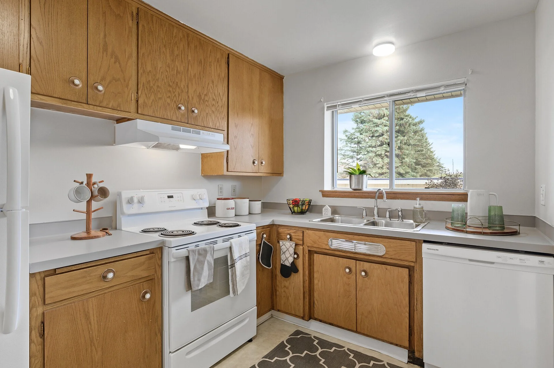 A kitchen with wooden cabinets, a white stove, a double sink by a window, and a dishwasher. There's a potted plant on the windowsill and decorative items on the counter at Imperial Heights in West St. Paul, MN