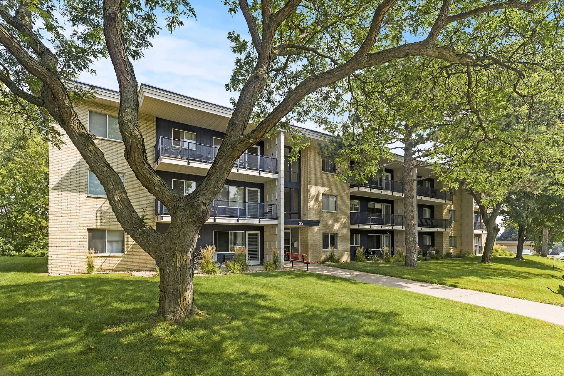 A multi-story residential building at Imperial Heights in West St. Paul, MN viewed through tall trees with green leaves and a well-maintained grassy lawn in the foreground.