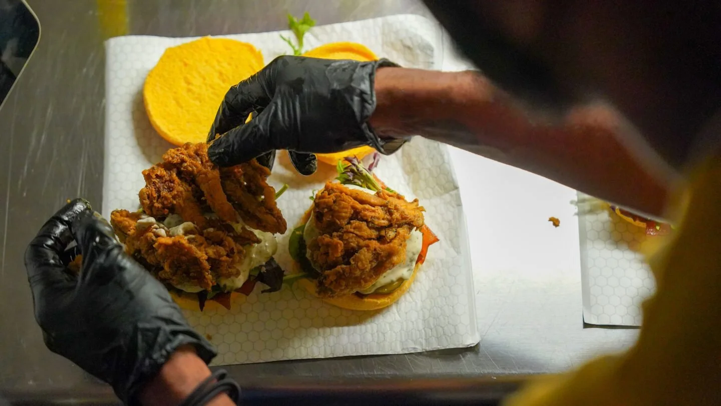 A person wearing black gloves assembling fried chicken sandwiches on a sheet of white patterned paper. The sandwiches contain fried chicken, lettuce, tomato, and mayonnaise, with a side of potato chips and sliced tomatoes in the background.
