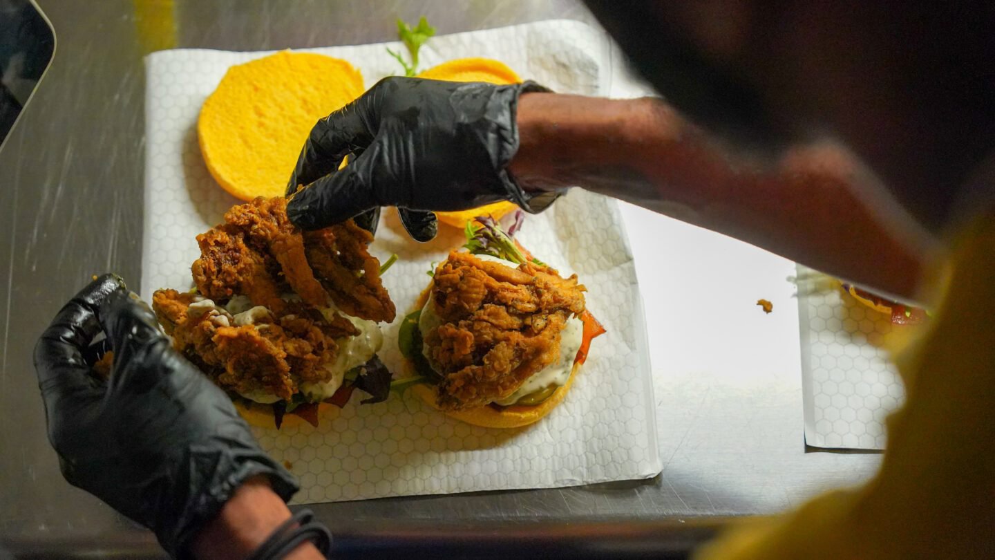 Person wearing black gloves assembling a fried chicken sandwich with lettuce, tomato, and sauce on a honey bun bun, with a side of potato chips in the background.