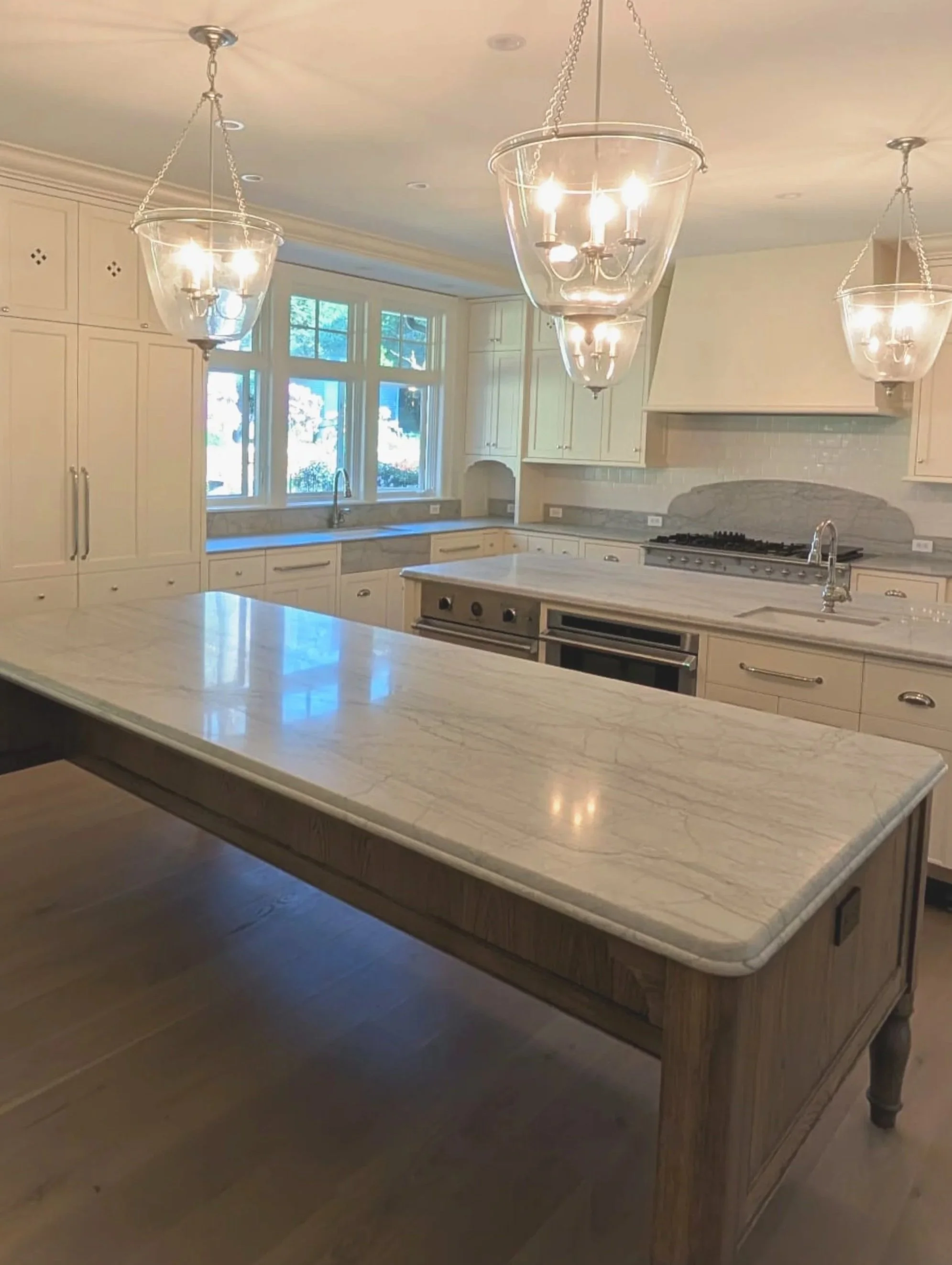 A white kitchen with marble countertops, hanging glass chandeliers, cabinetry, a large window, and a stove.