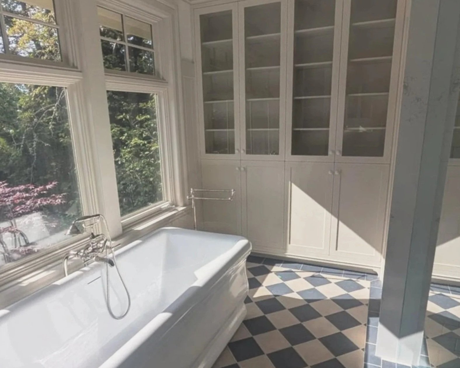 A bathroom with a built-in white bathtub next to large windows, white cabinets with glass doors, and a patterned tile floor.