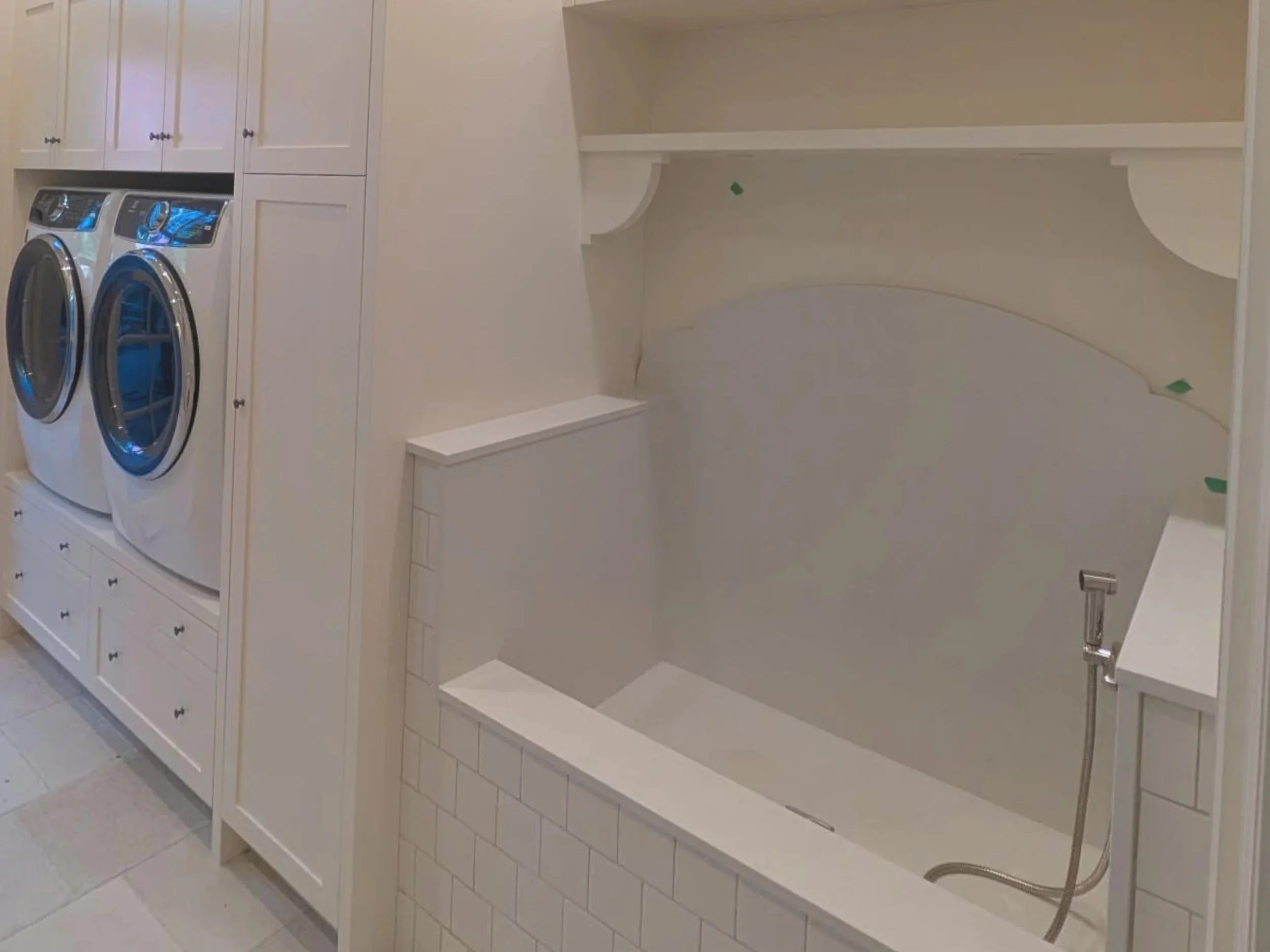 White laundry room with front-loading washing machine and dryer, white cabinets above, and a built-in shelf with decorative molding, corner space, and tiled flooring.