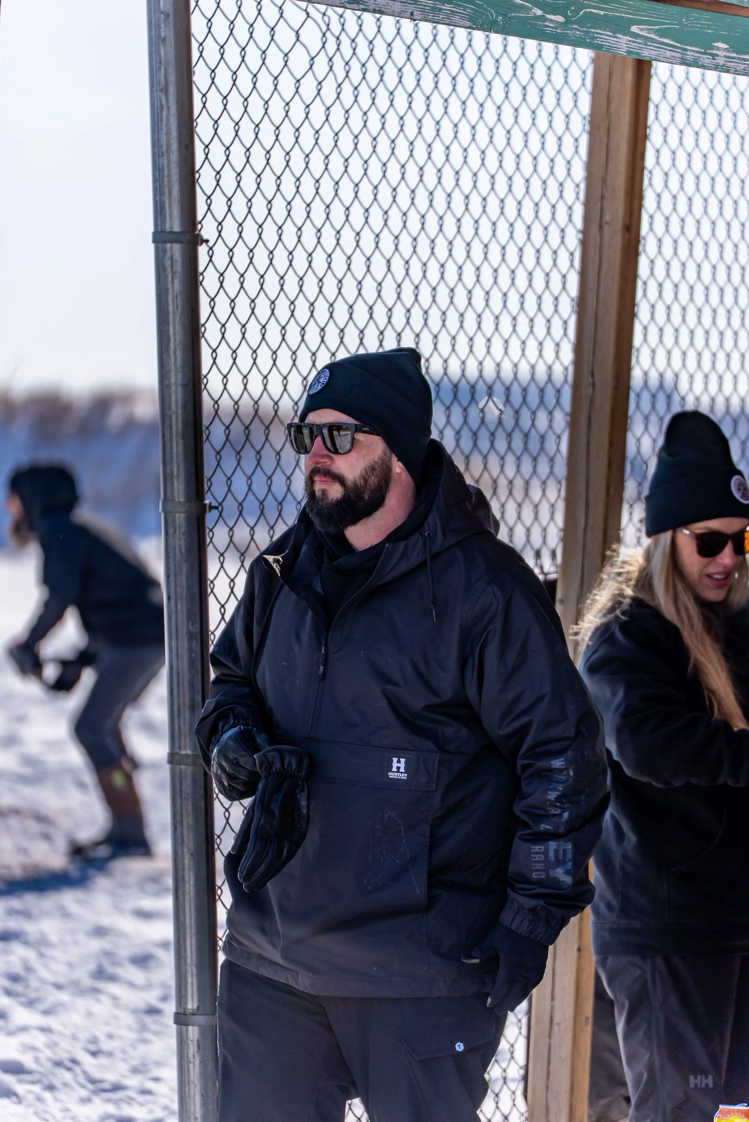 A man wearing sunglasses, a black beanie, and a black jacket standing near a chain-link fence outdoors in a snowy environment during daytime.