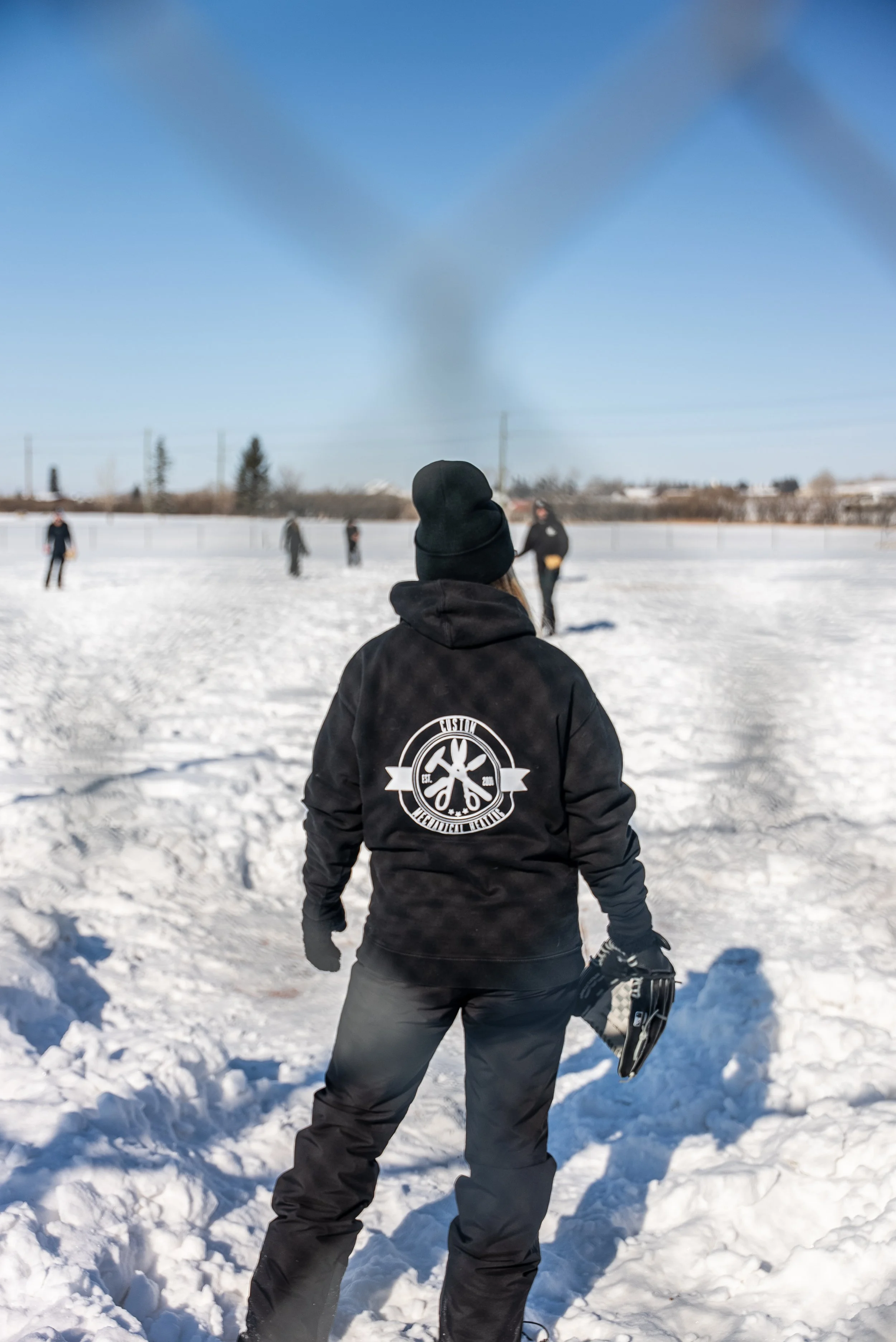 A person dressed in black winter clothing holding a hockey stick and glove, standing in snow-covered ground during daytime, with other people in the background playing hockey or standing in the snow.