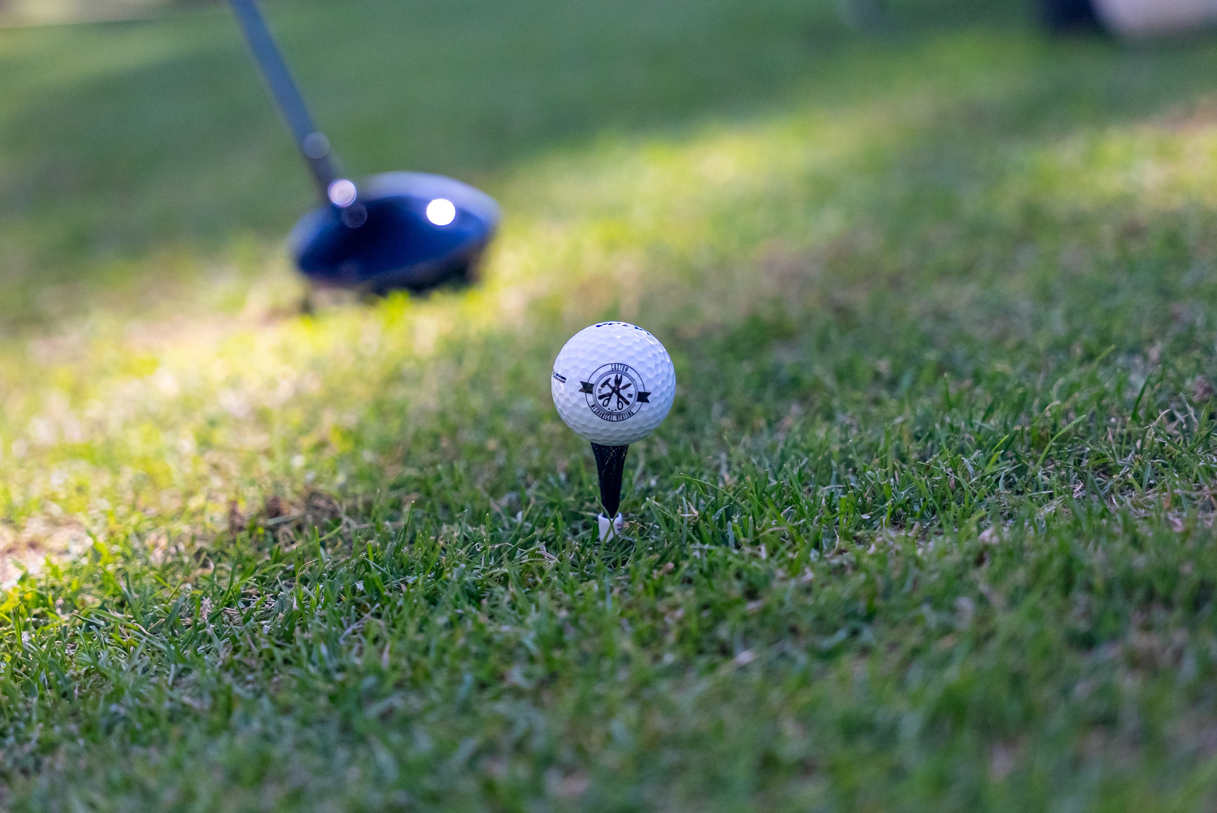 Close-up of a golf ball on a tee on the grass with a golf club approaching in the background.