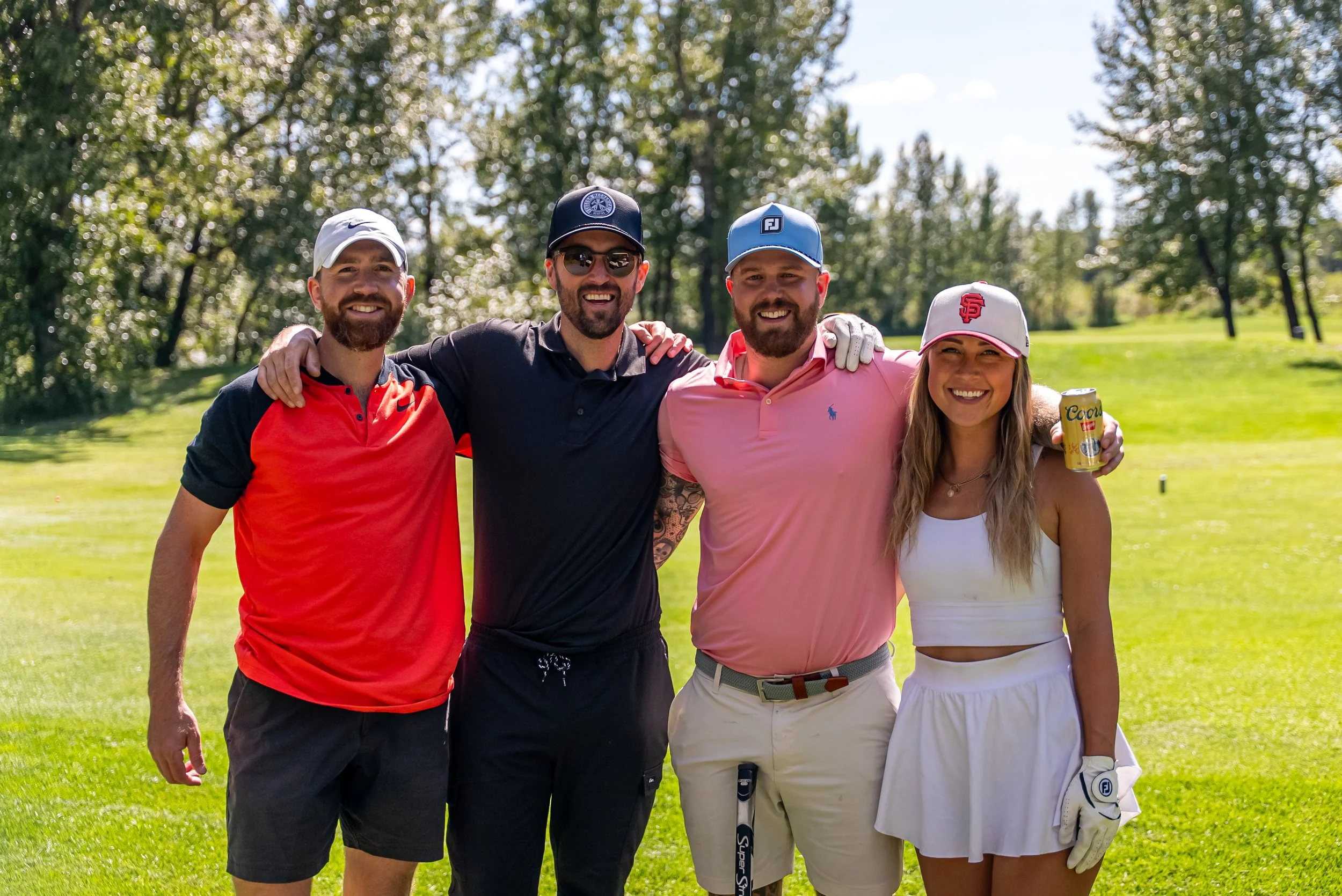 Group of four friends smiling and standing together outdoors on a golf course with trees in the background. They are wearing casual summer clothing and hats, with one woman holding a beer can.