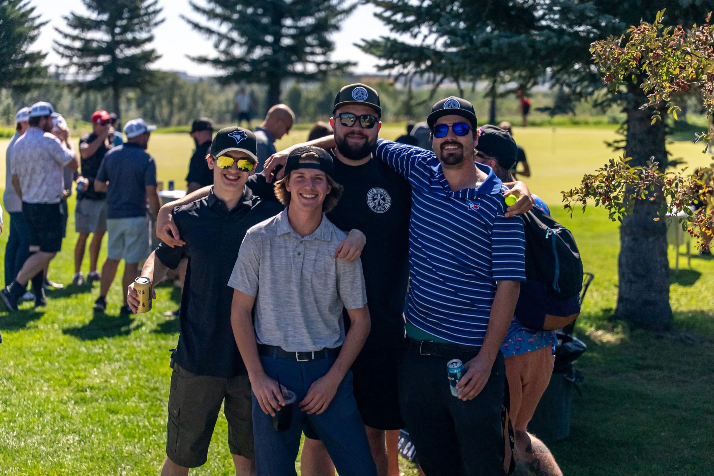Group of four friends standing outdoors in a grassy area, smiling and posing for a photo on a sunny day, with other people in the background.