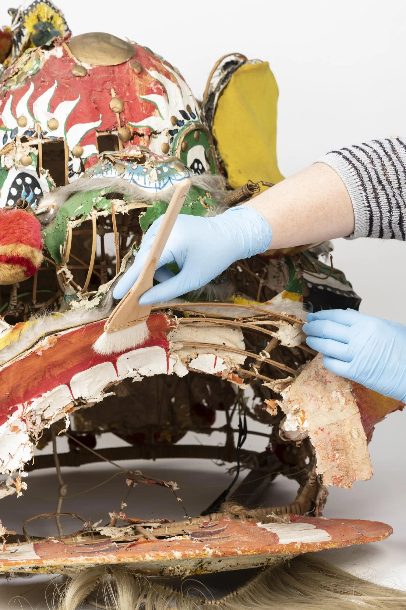 Close-up of a damaged and weathered doll, with a painted face and long hair, wearing a colorful, torn costume. Someone in a striped shirt with blue gloves is carefully working on it with tools.