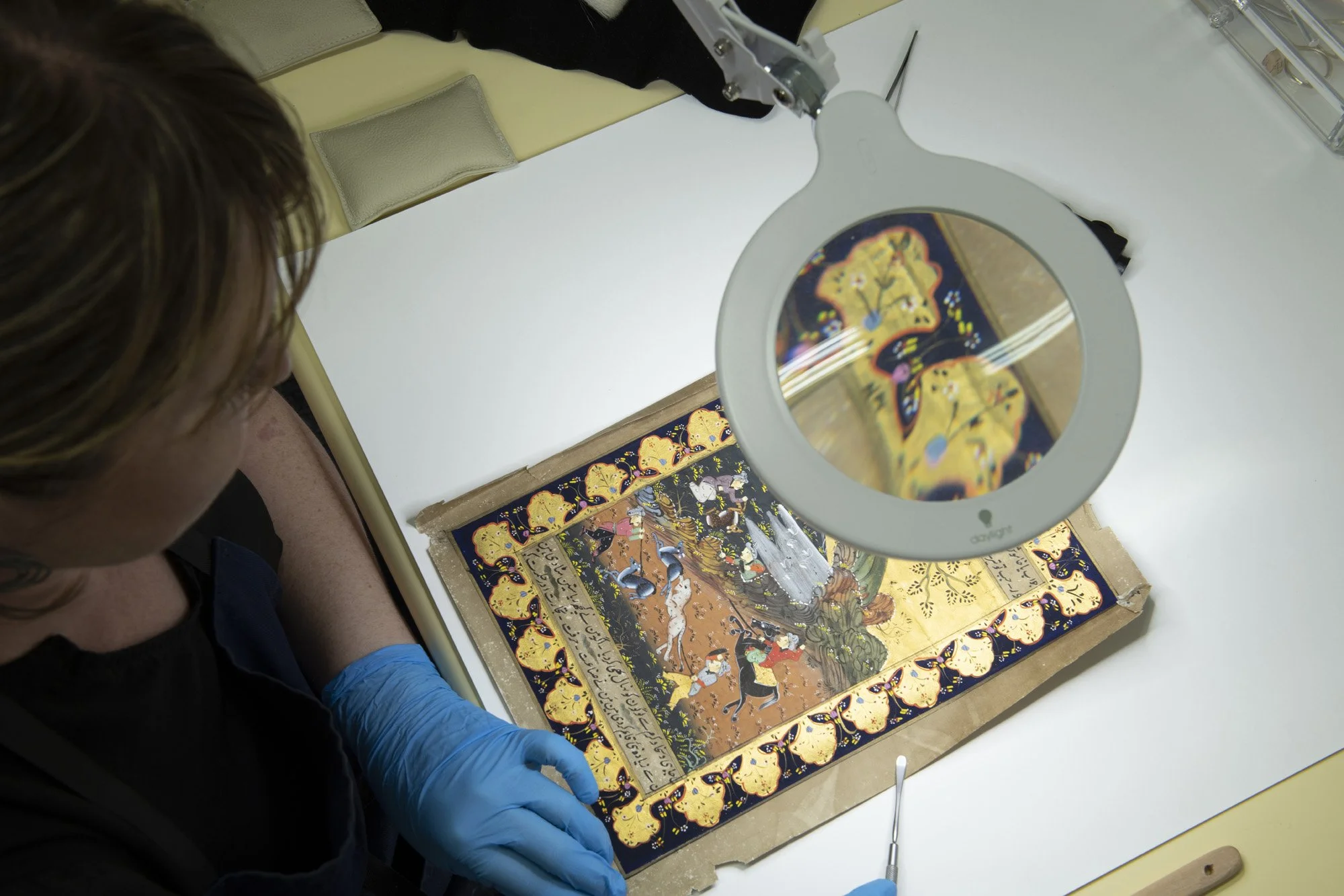 A person wearing blue gloves examining an ancient religious manuscript or painting under a magnifying glass in a museum or conservation lab.