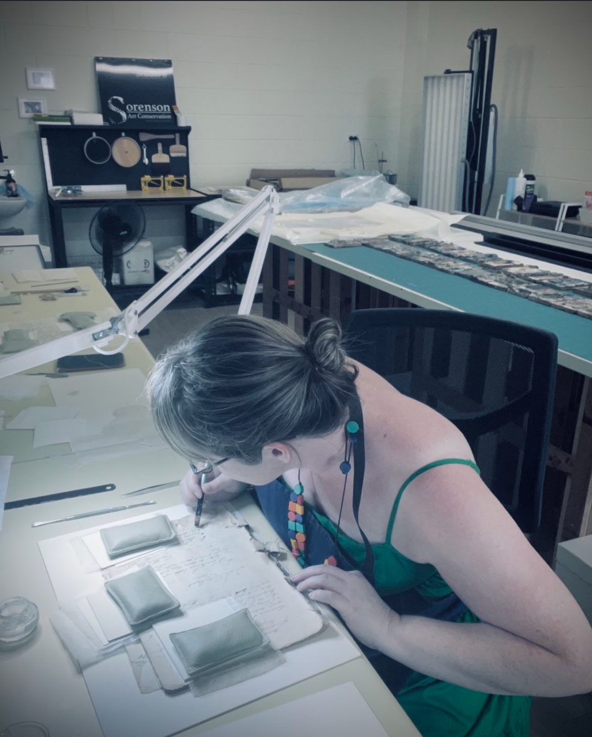 A woman working at a desk in a laboratory or workshop, examining and working on documents or samples surrounded by various tools and materials.