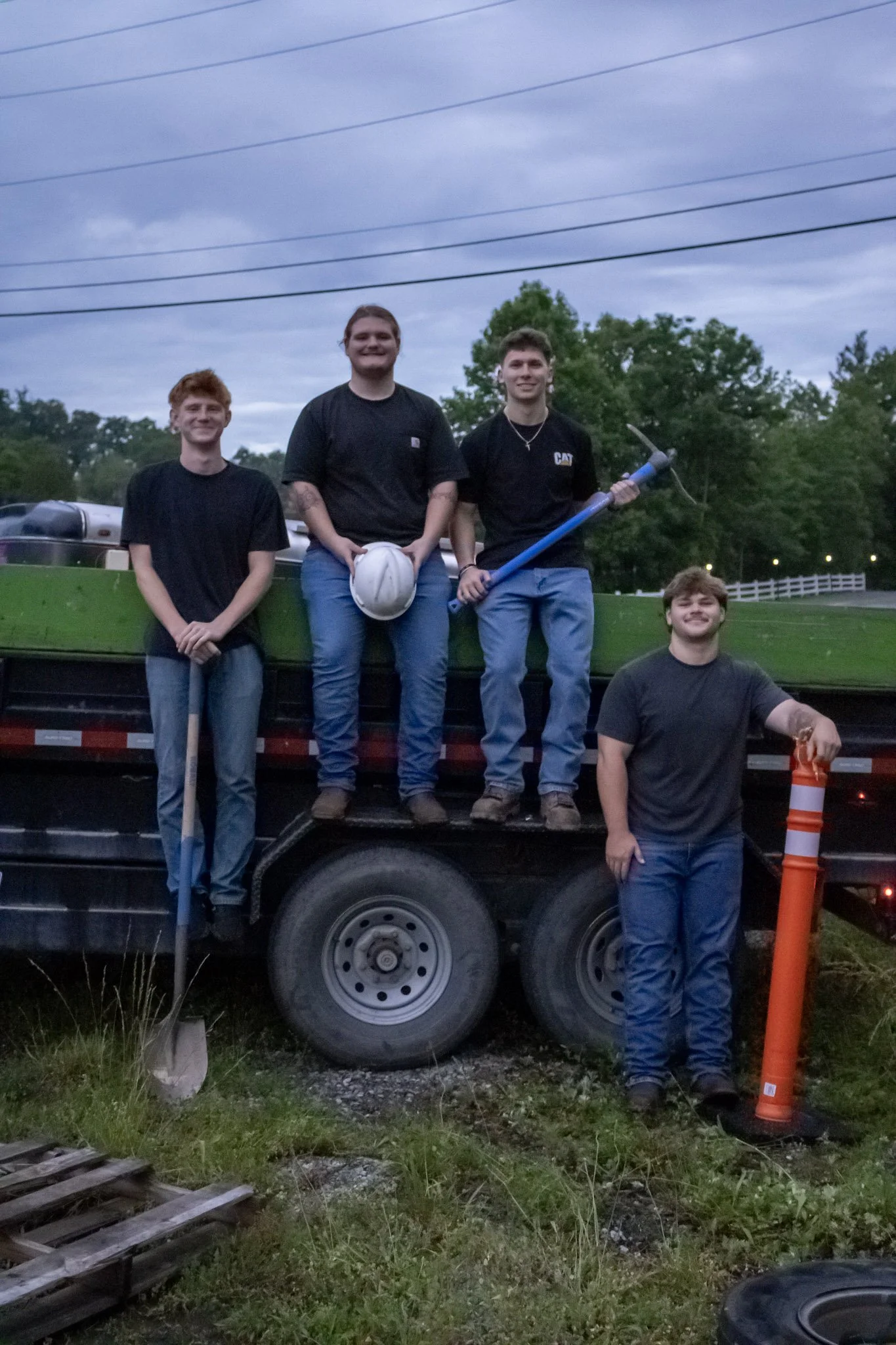 Four Junktion employees standing on a truck bed outdoors during evening, with trees and a cloudy sky in the background. One is holding a large pickaxe, another has a white helmet, and they are smiling.
