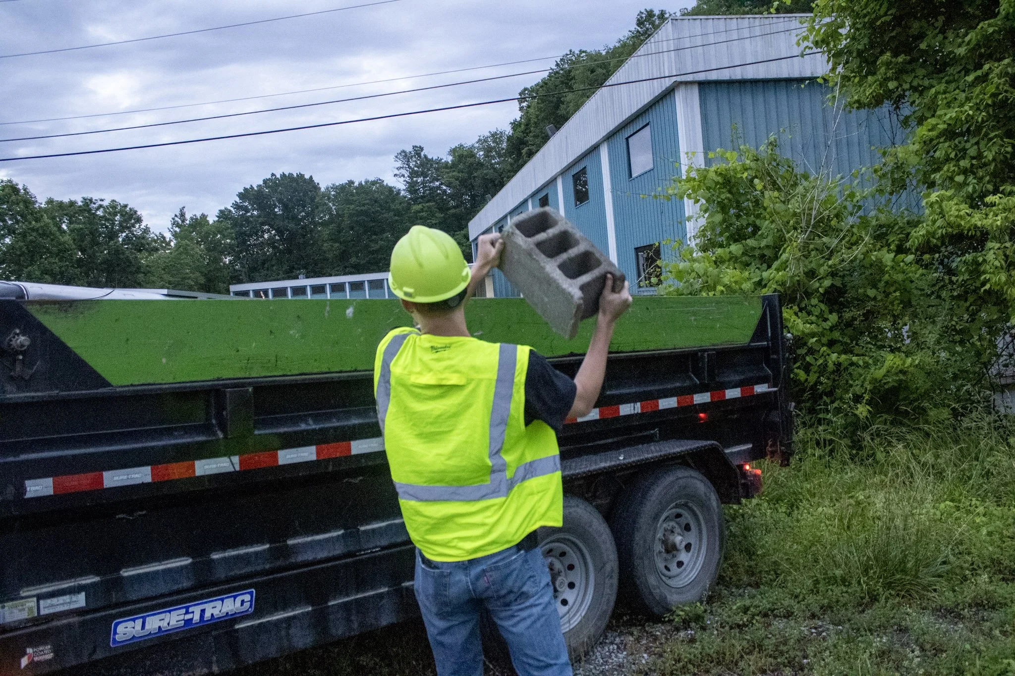 A Junktion employee loading a cinder block onto a trailer during daytime with overcast skies.