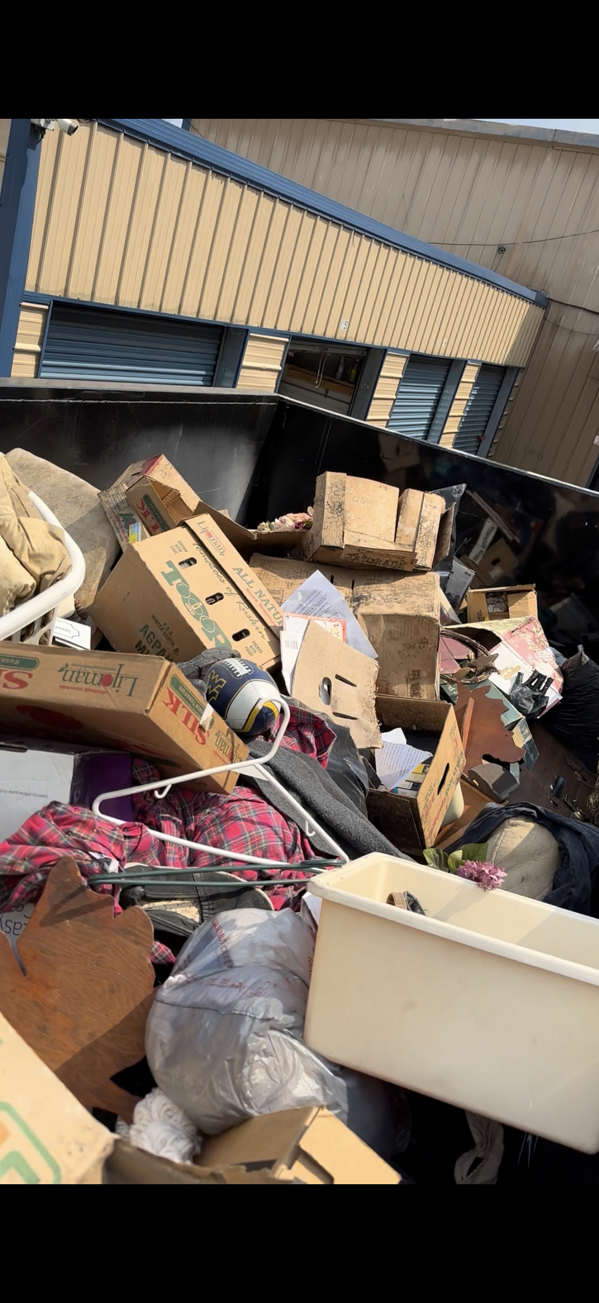 A large pile of trash and discarded items in a black dumpster, with cardboard boxes, plastic bags, clothing, and other debris against the background of a building with metal siding and windows.