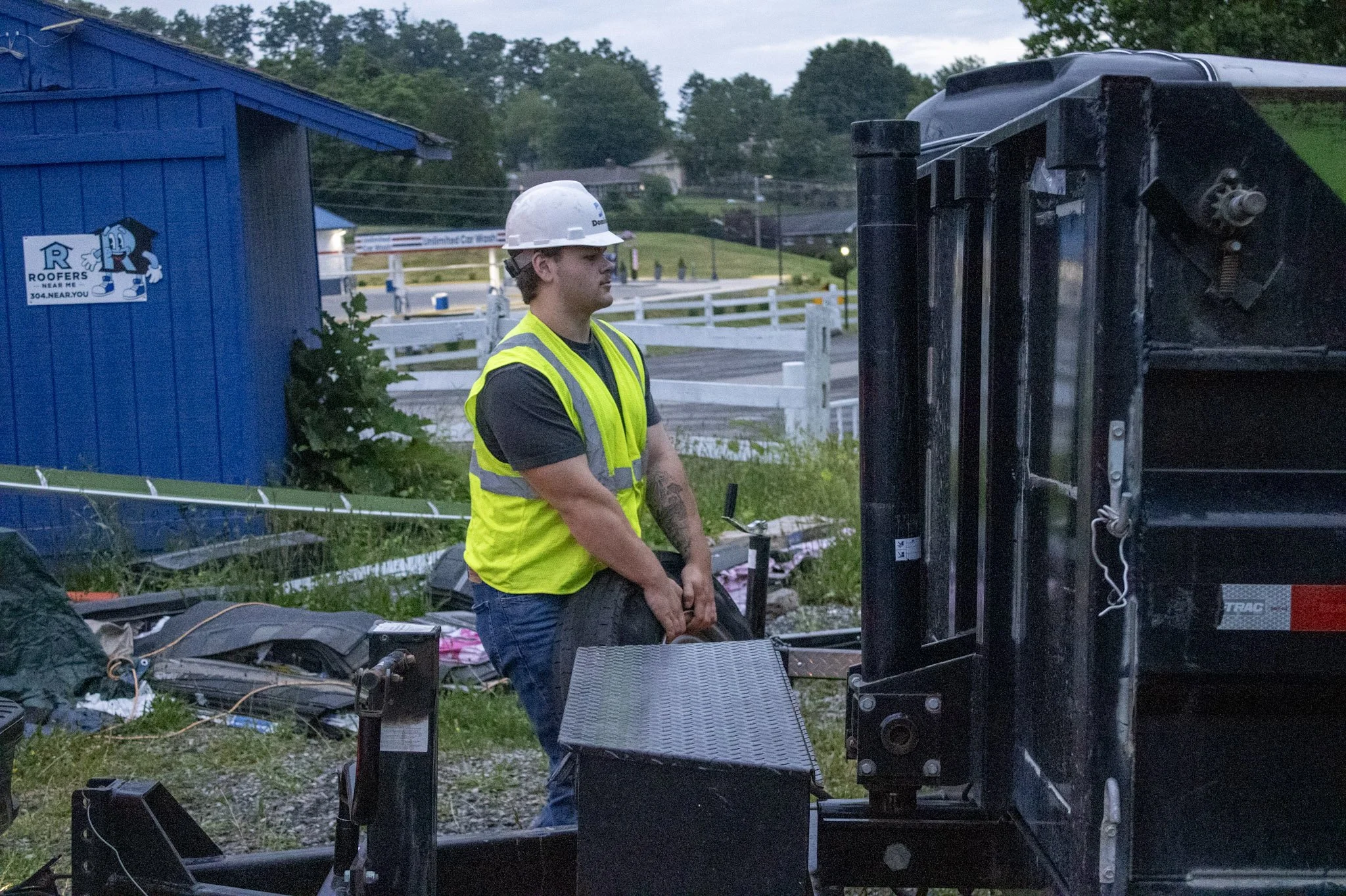 A young man wearing a yellow safety vest and white hard hat stands outdoors near a black vehicle or equipment.