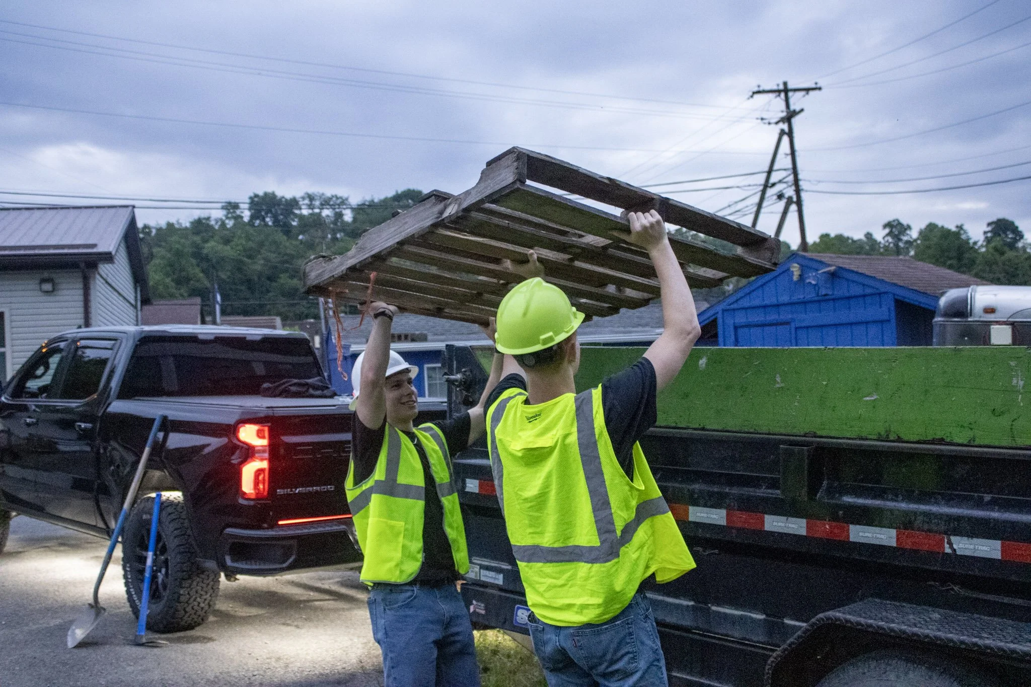 Two Junktion Employees wearing yellow safety vests and hard hats are lifting a wooden pallet in a parking lot during overcast weather.