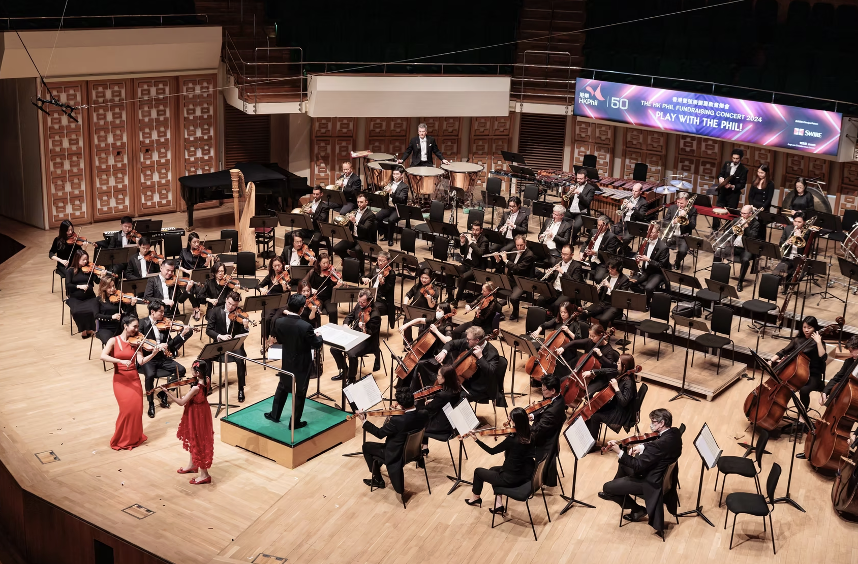 An orchestra performing on stage with a conductor, two women in red dresses are performing in front of the orchestra, a digital banner reads 'HK Phil Fundraising Concert 2024, Play with the Phil!'
