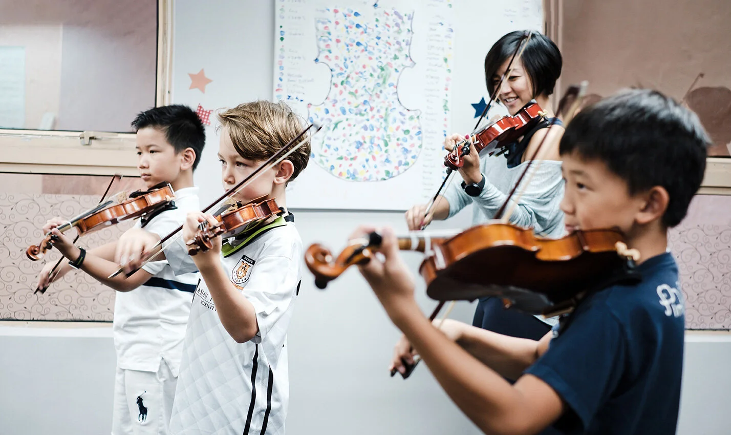 Four children playing violins in a classroom, with a colorful map on the wall behind them.