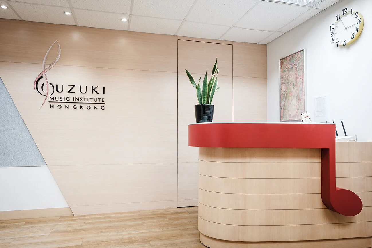 Reception area of Ouzuki Music Institute in Hong Kong with a wooden desk, a potted plant, a large musical note sculpture on the desk, and a clock on the wall showing 2:43.