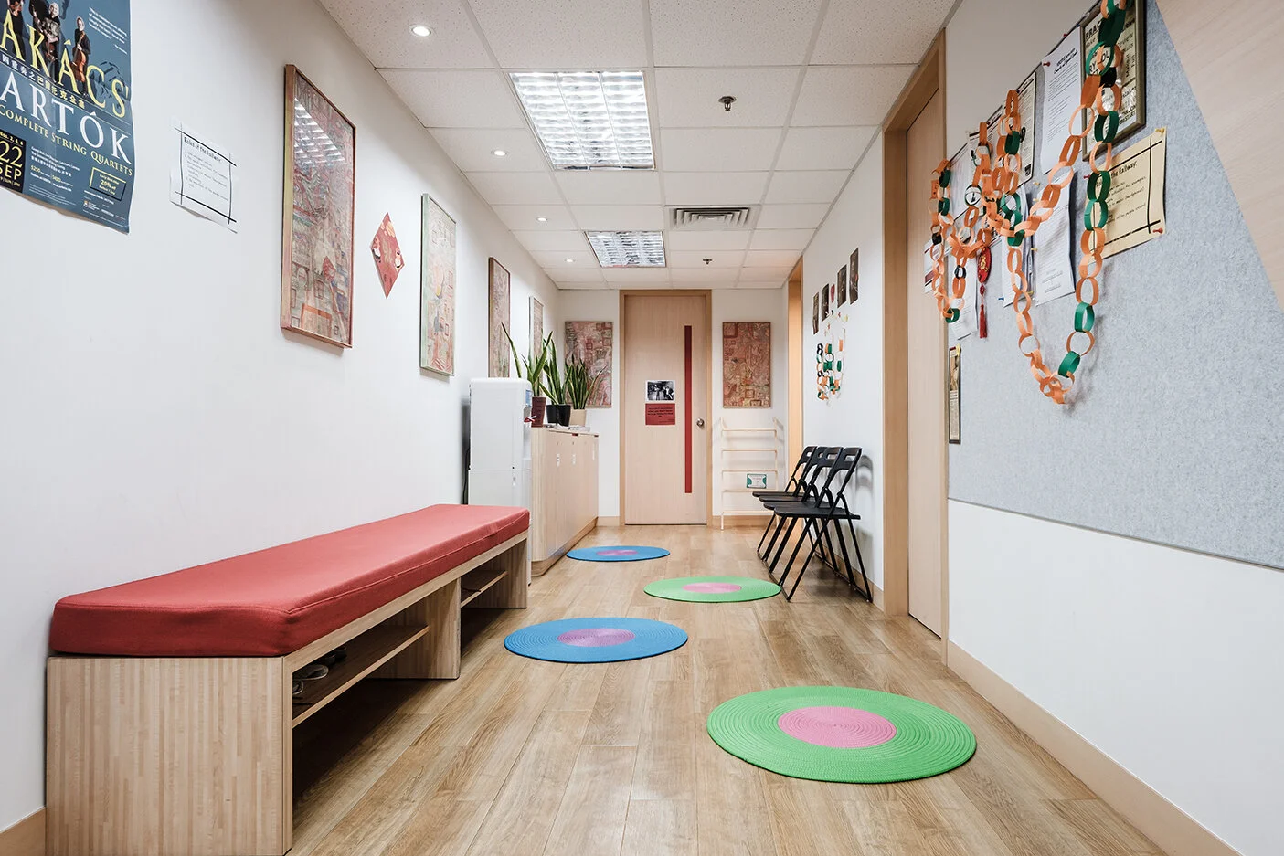 Empty waiting room or hallway with black chairs, colorful circular rugs, a red bench, framed posters on white walls, a bulletin board, and some decorations.