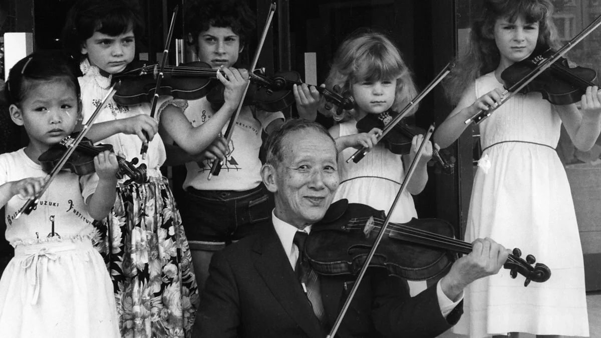 A group of young girls and Dr. Shinichi Suzuki playing violins, with the man sitting and the girls standing behind him, in black and white.