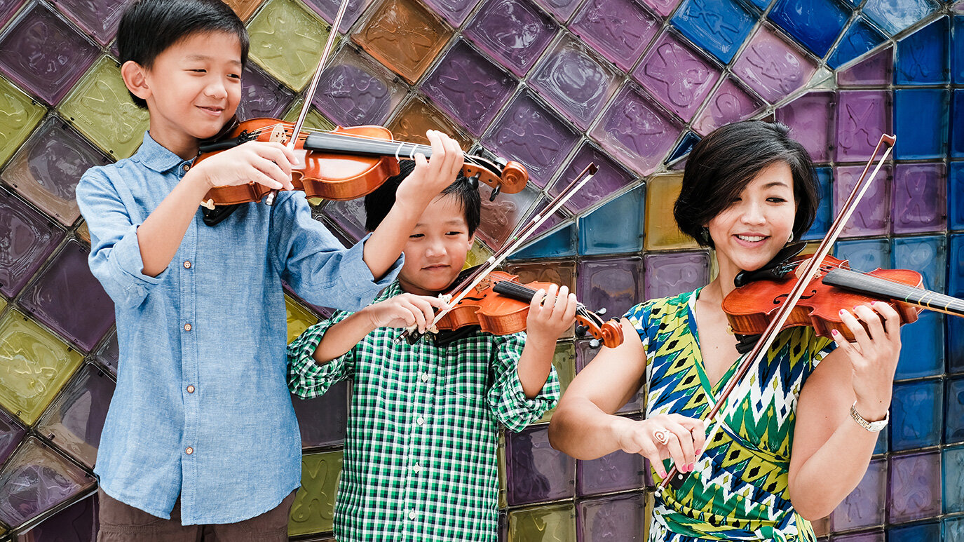 Three children and a woman playing violins outdoors against a colorful tiled wall.