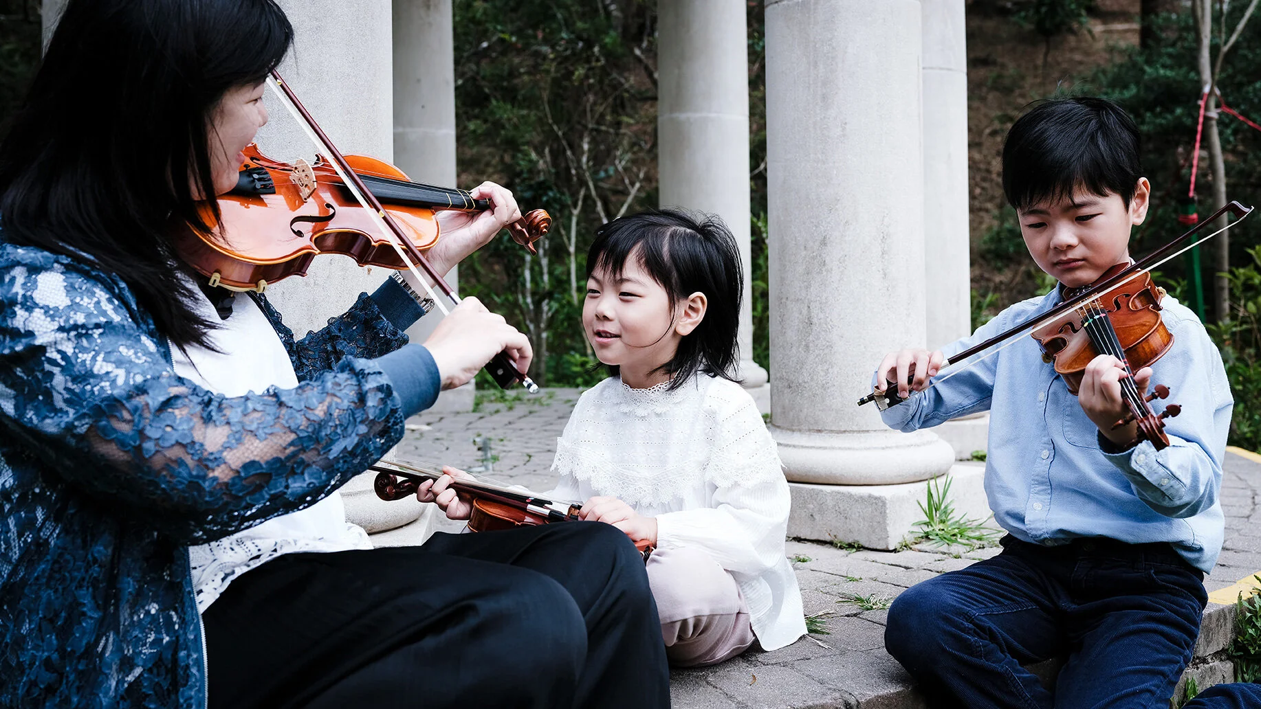 A woman and two children playing violins outdoors near white columns and greenery.