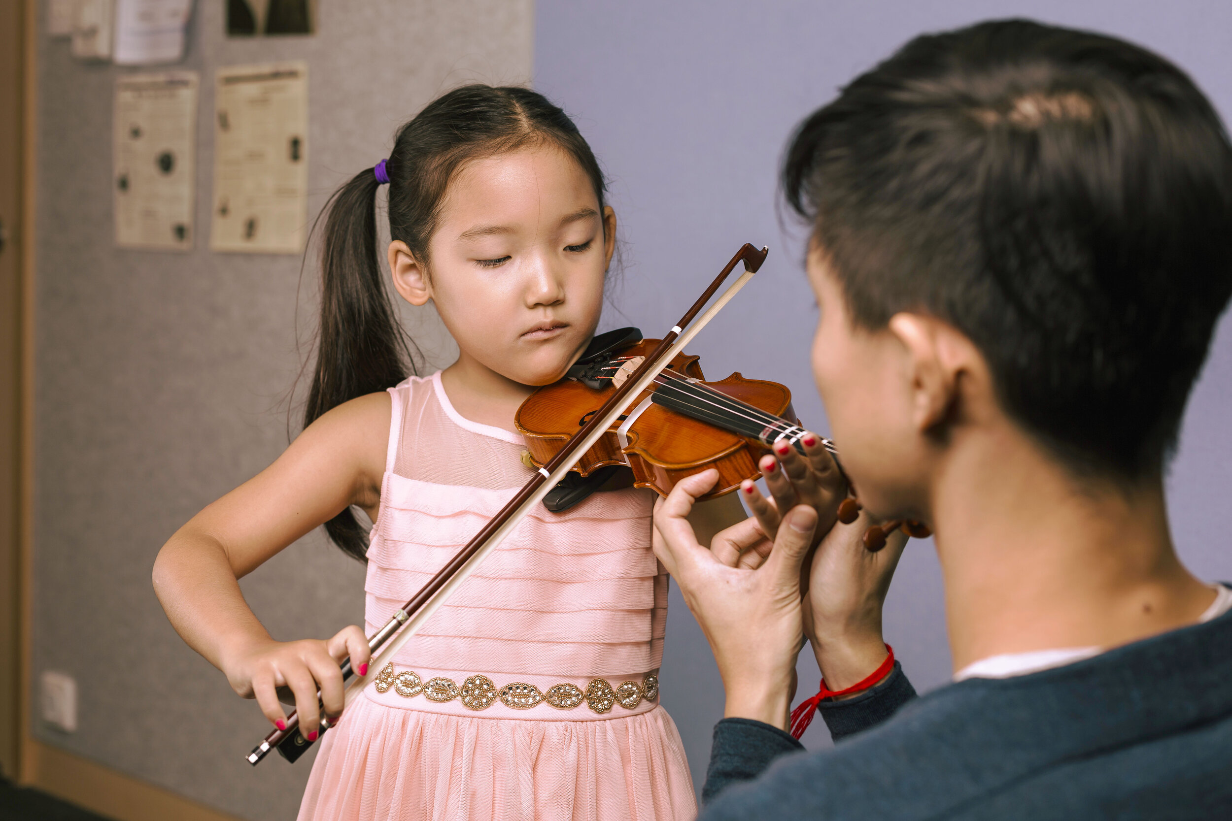 A young girl with long dark hair in two ponytails, wearing a pink dress, is holding a violin. A teacher is assisting her with the violin, holding it in place near her neck. They are in a room with posters and papers on the wall.