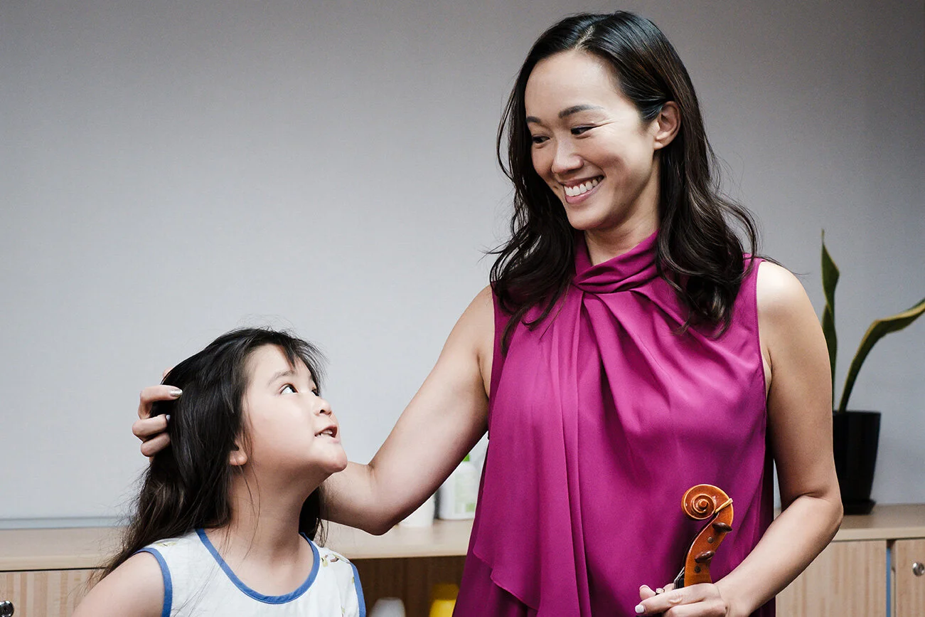 A woman in a magenta sleeveless top holding a violin smiles at a young girl with long dark hair and a blue and white top, as the woman gently pats the girl's head.