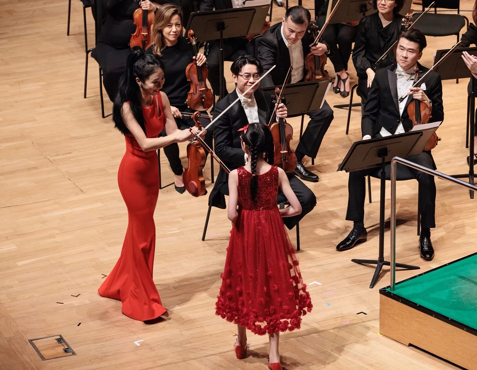 Orchestra members on stage, with two women in red dresses in the front, one holding a violin and the other a microphone, and several men in tuxedos holding violins, one with a music stand in front of him. A young girl in a red dress with a red bow in her hair is in the foreground.