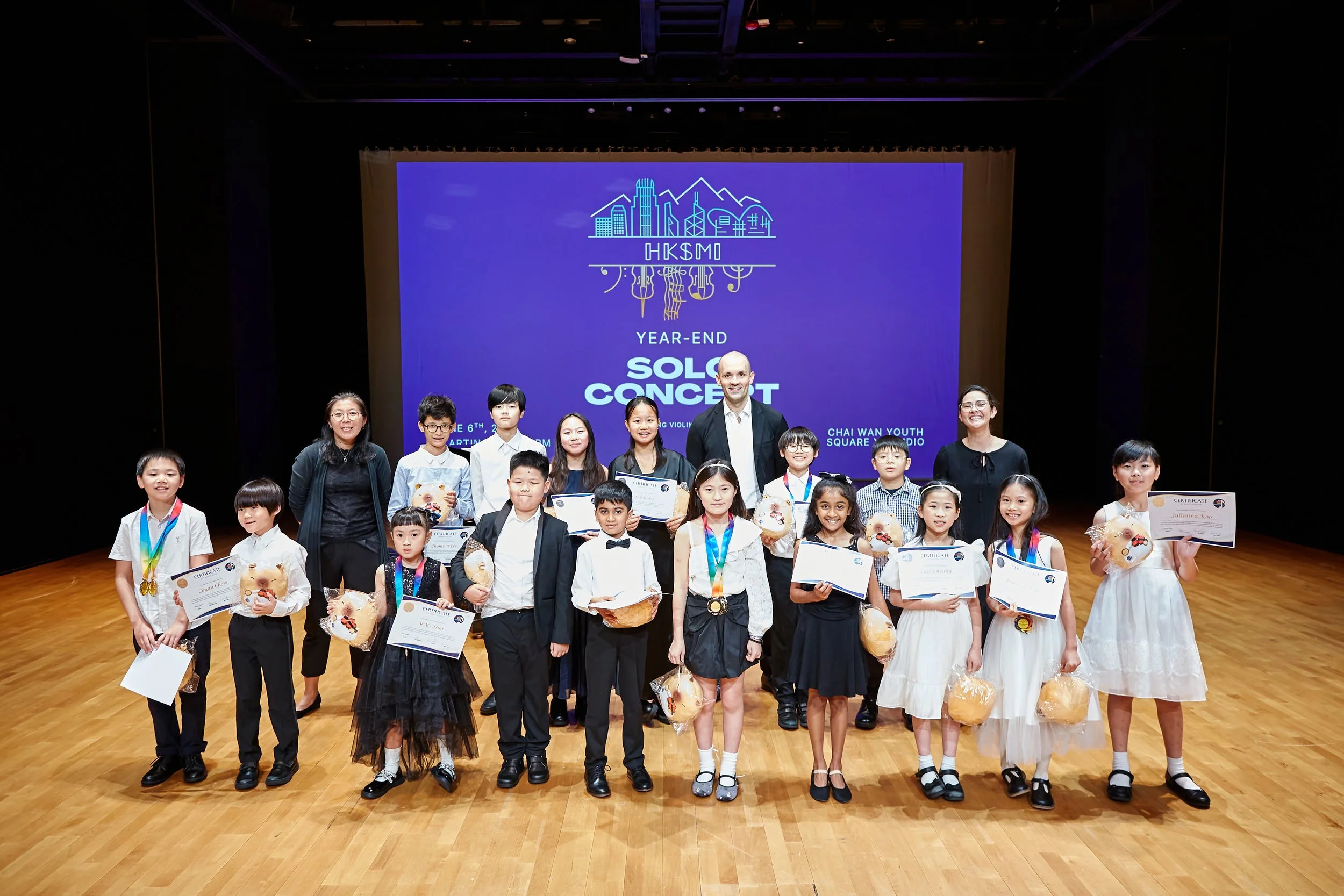 Group of children and adults on stage holding certificates and prizes at a year-end event, with a large screen displaying the event information behind them.