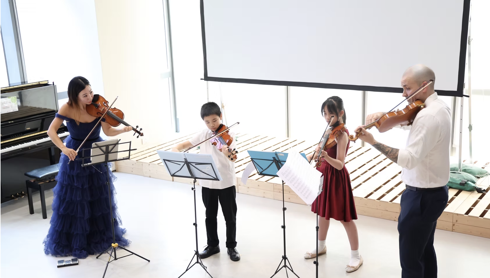 Young Virtuosi Program: Four musicians playing violins in a bright room. Two women and two children, all reading sheet music.