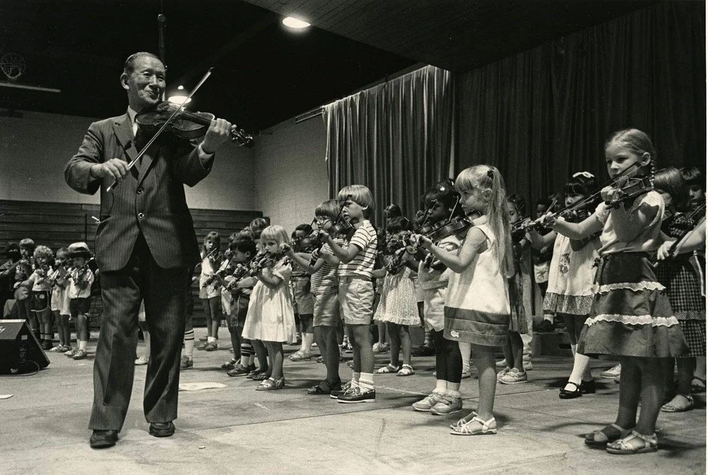 Dr.Shinichi Suzuki plays the violin on stage while a group of children also play violins, practicing in a school gymnasium or auditorium with dark curtains and a wooden floor.